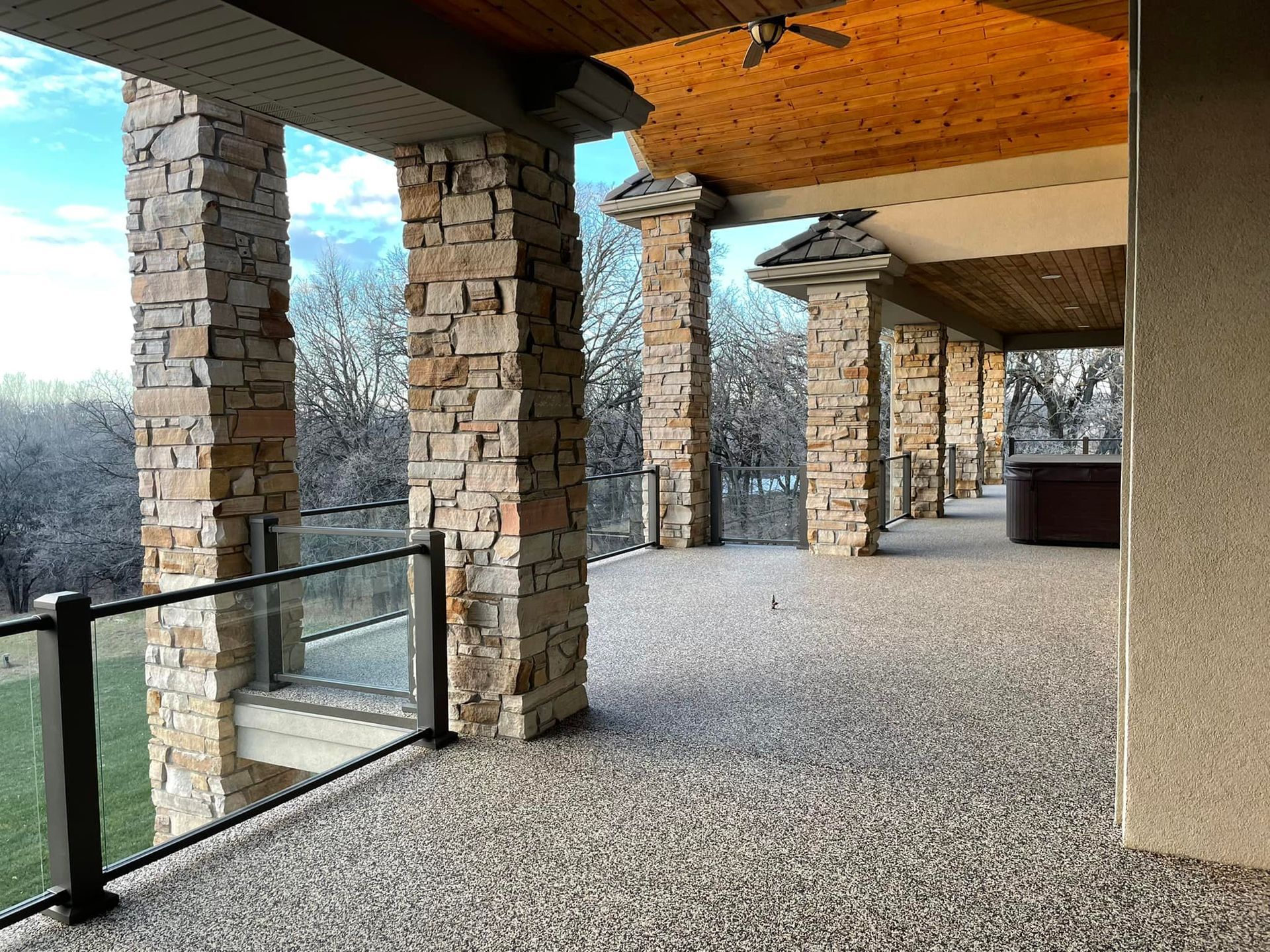 A spacious, covered outdoor patio featuring stone columns, a textured pebble floor, and glass railings overlooking trees