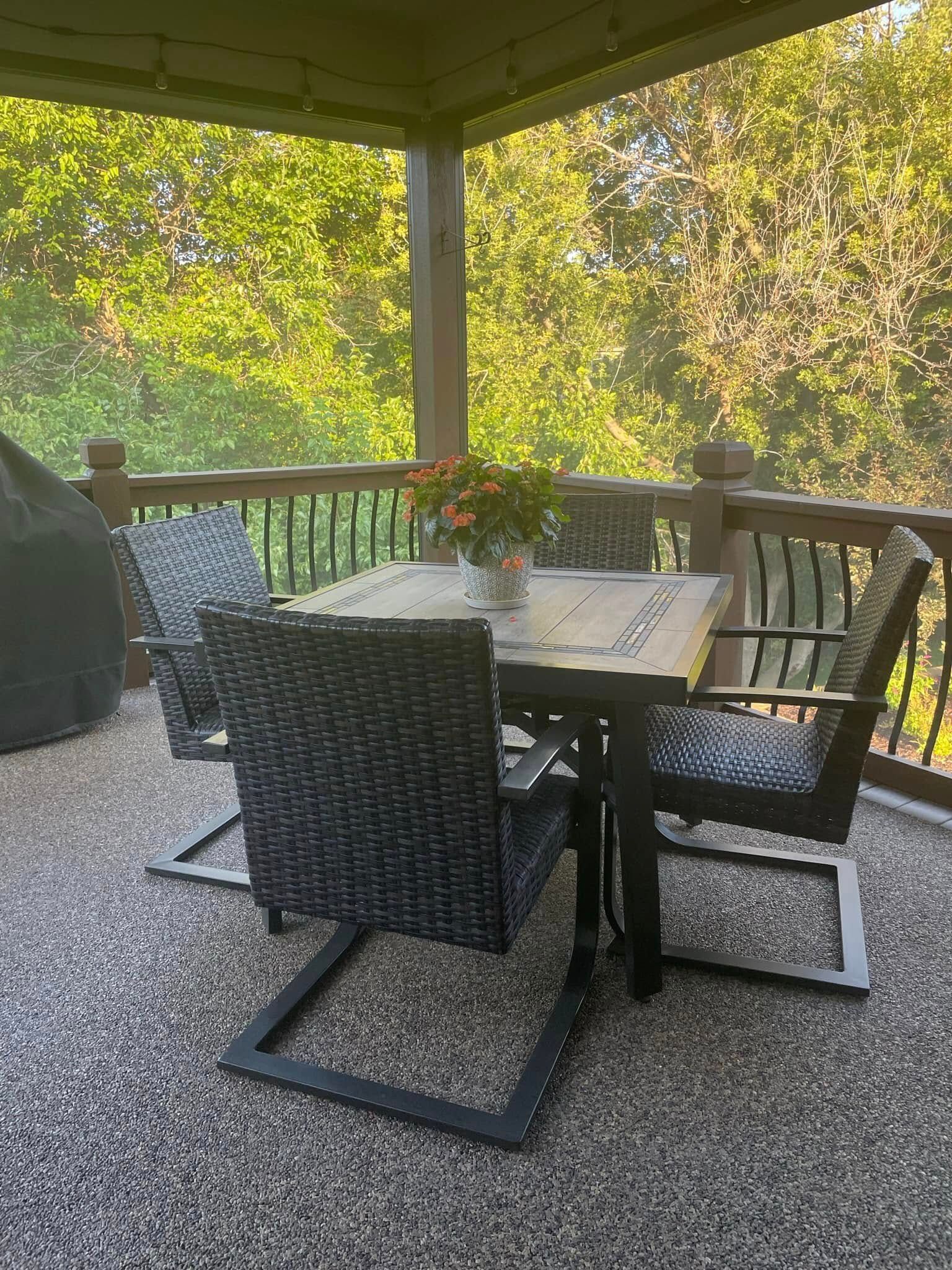 A patio set with a square table and four black wicker chairs on a textured stone balcony overlooking trees.