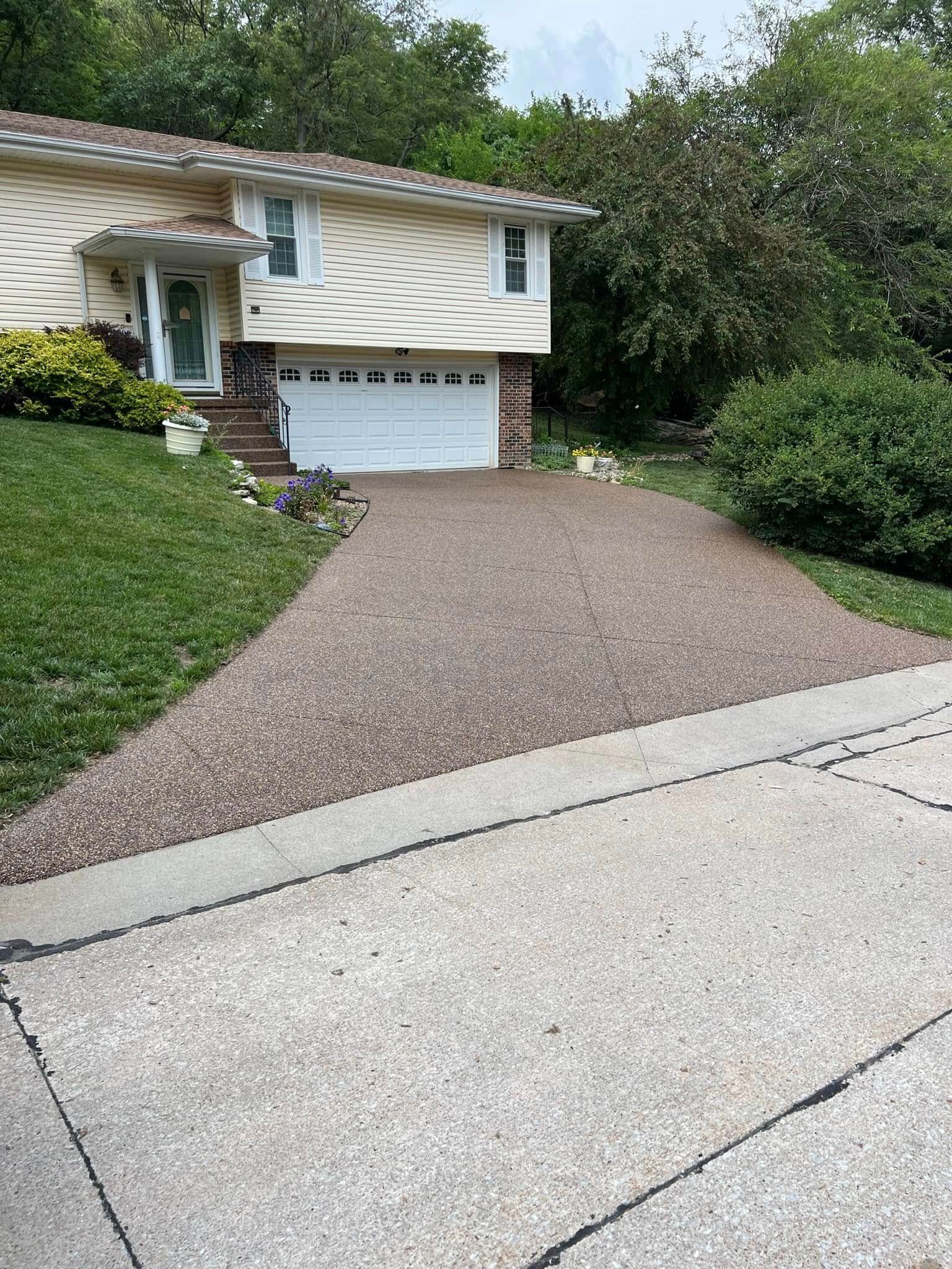 A beige split-level house with a white garage door and an exposed-aggregate driveway, surrounded by trees and greenery