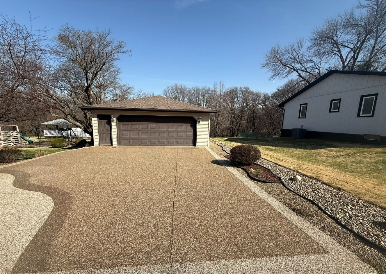 A gravel driveway leads to a detached garage with a dark door, situated next to a white house on a sunny day