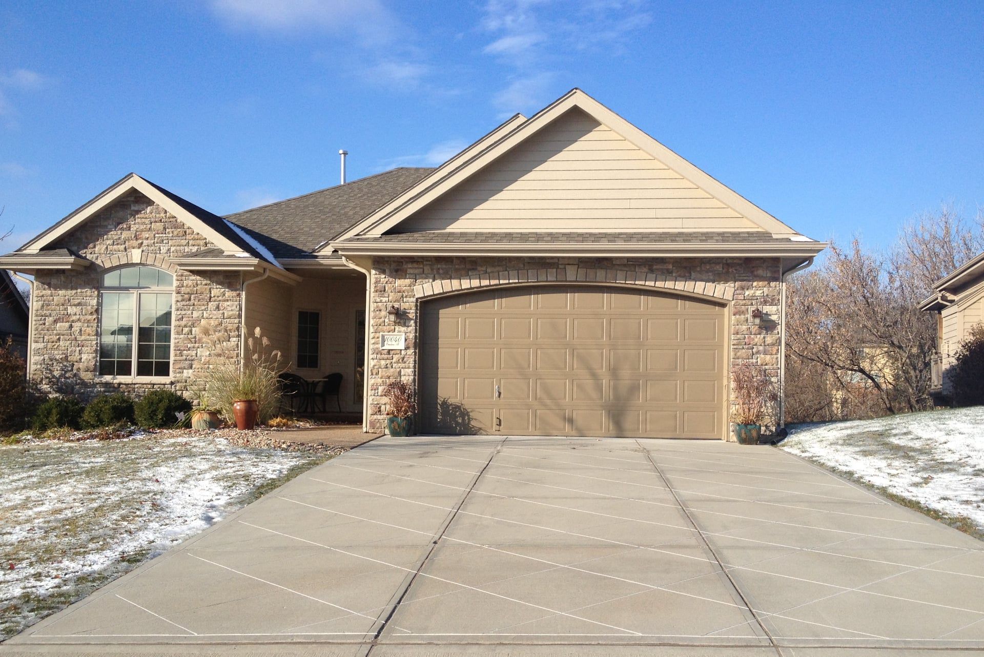 A single-story tan stone house with a double garage and a concrete driveway on a sunny day with patches of snow.