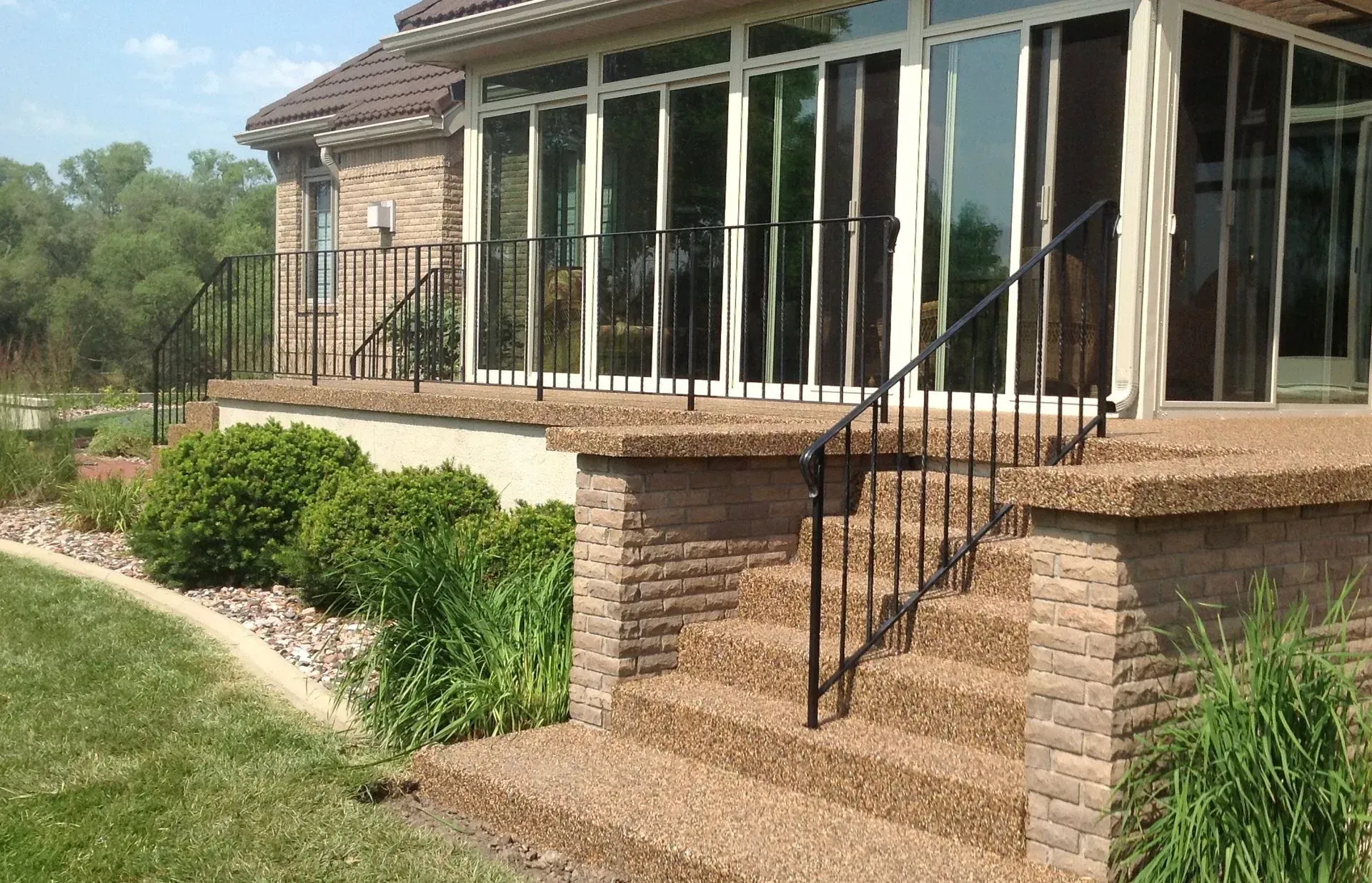 A brick patio with a glass sunroom, black metal railings, and stone stairs leading down to a green lawn.