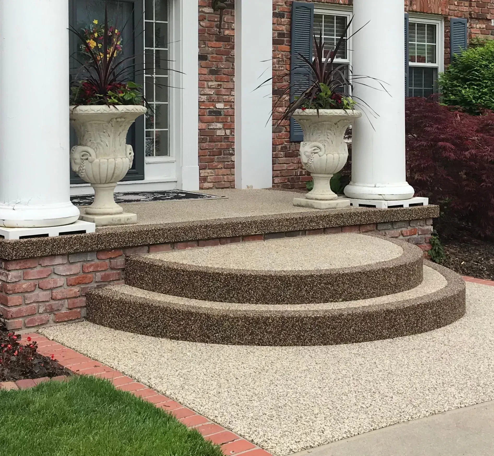 A front entryway with two semi-circular textured concrete steps, brick foundation, and two stone urns with tall plants.