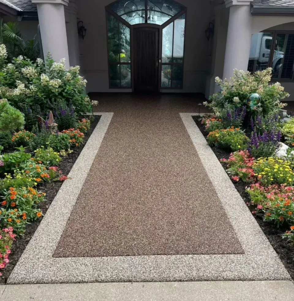 A walkway with a brown aggregate center and light stone border leading to a dark arched front door framed by gardens.