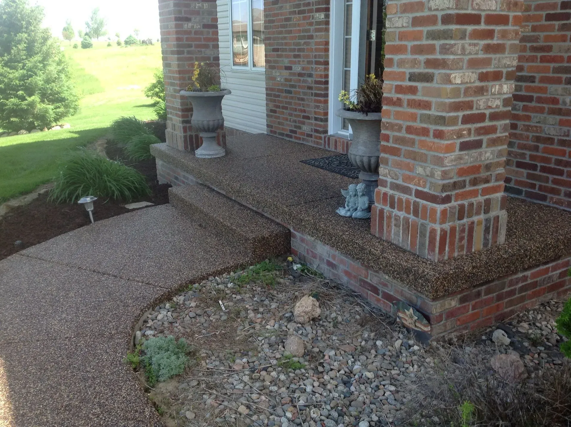A brick house entryway features a textured pea-gravel walkway leading up to a porch with two stone urn planters.