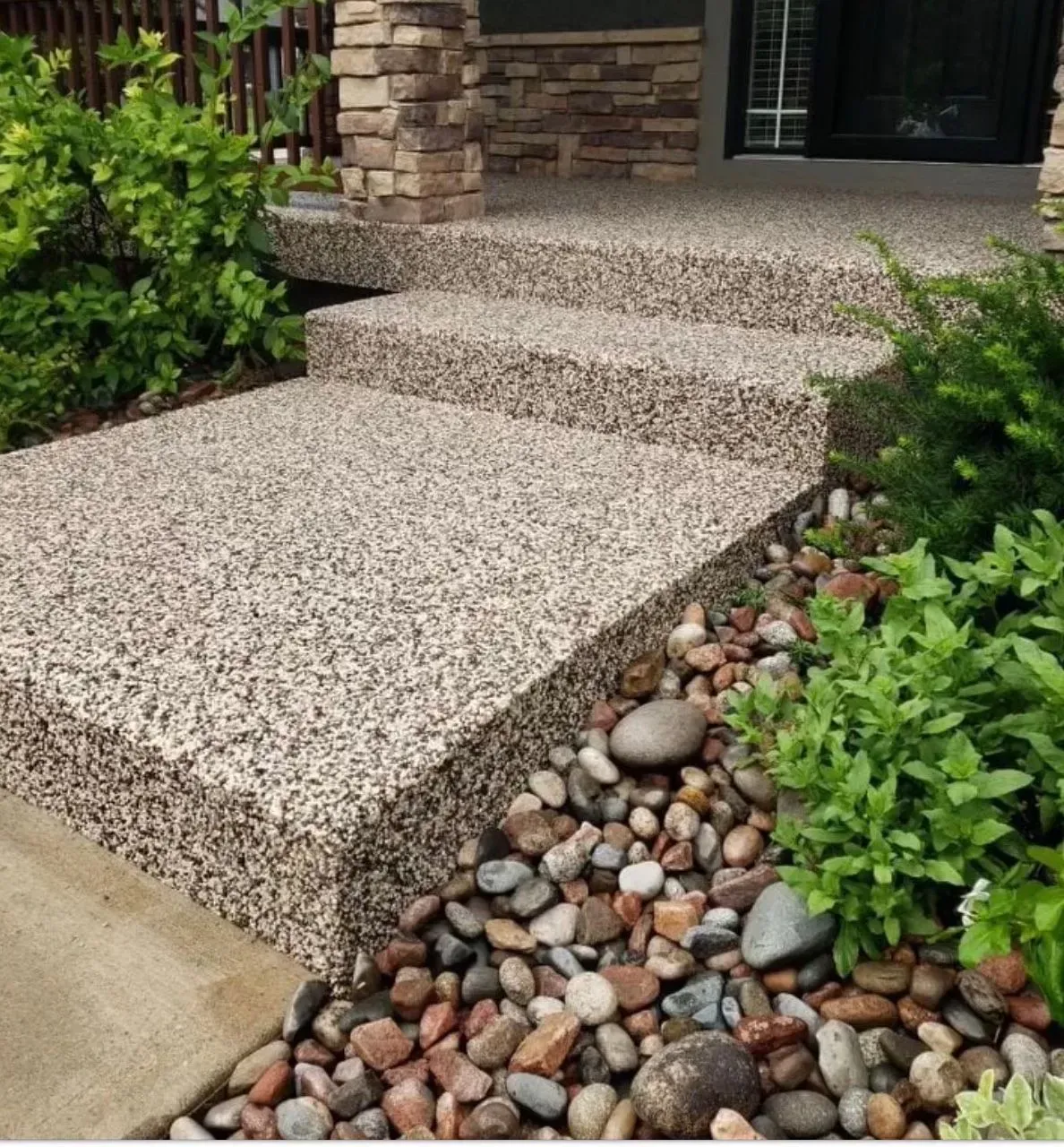 A small set of light-colored, textured stone-finish outdoor stairs leading to a home entrance, flanked by garden bushes.