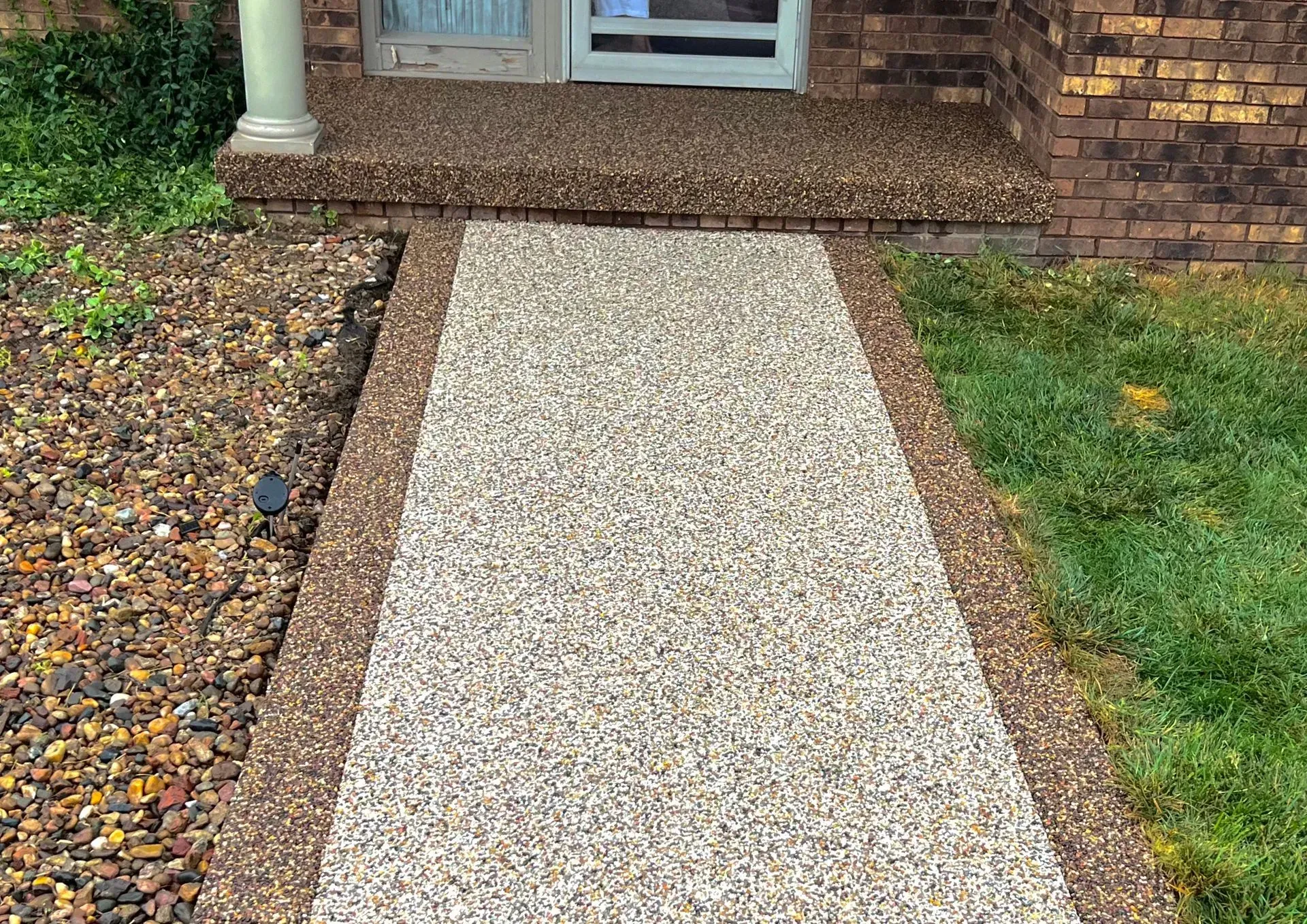 A light-colored stone walkway framed by dark brown stone borders, leading to a brick house entrance.