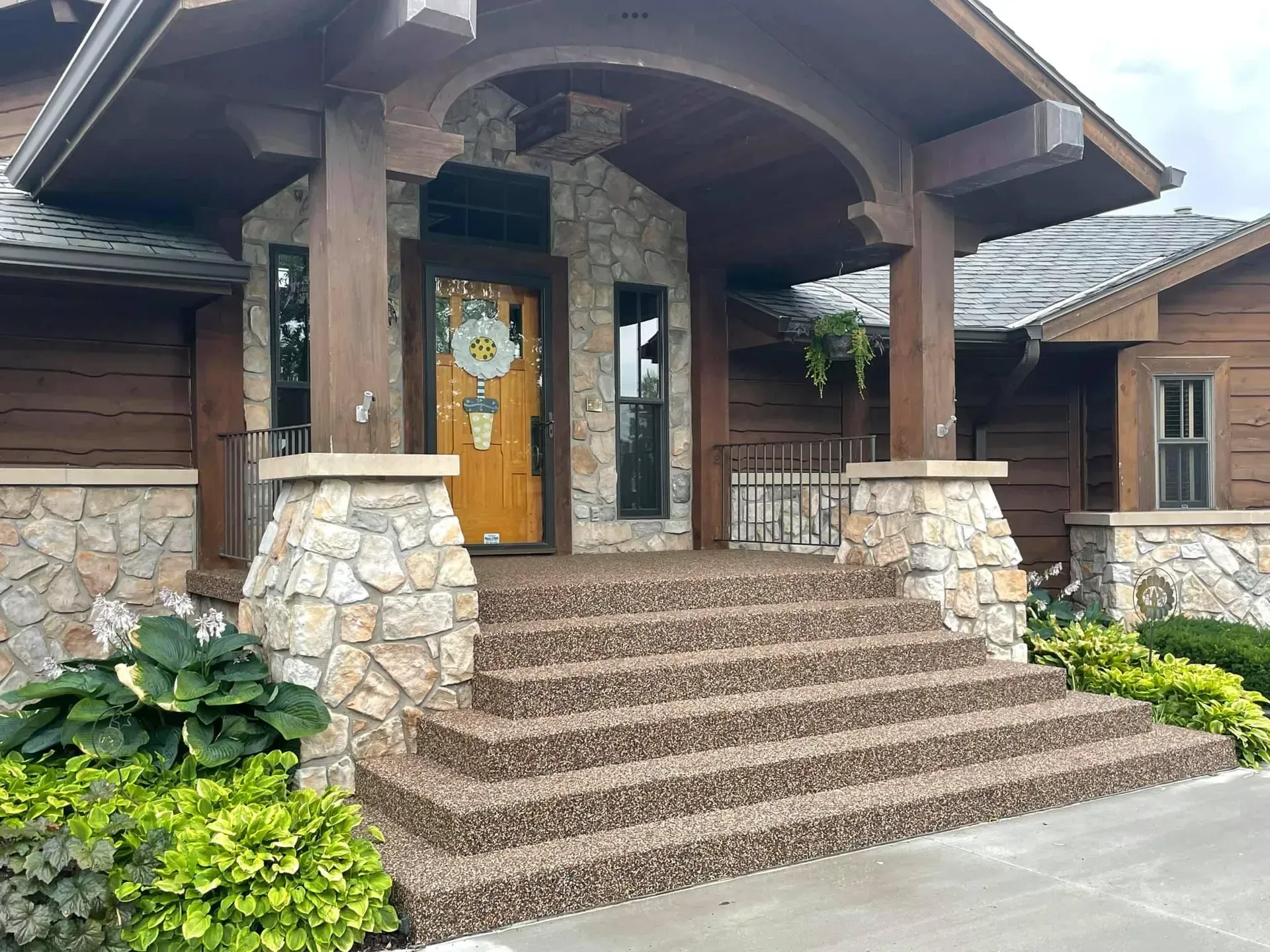 Rustic home entrance with stone walls, wooden beams, brown pebble stairs, and a wooden door with a floral wreath.