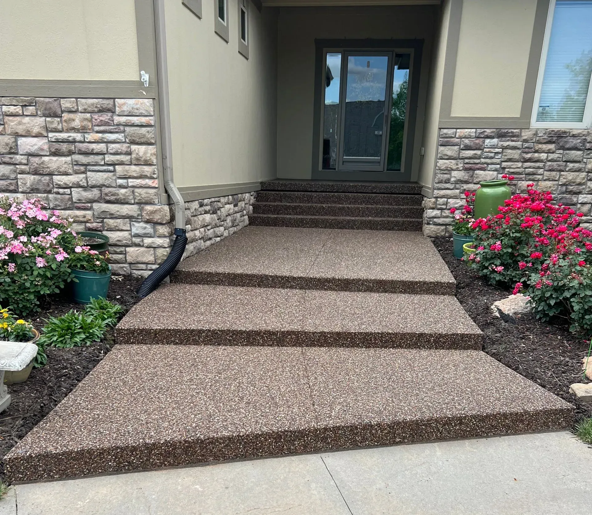 Concrete stairs with textured brown aggregate leading up to a house entrance with stone siding and pink flower beds.