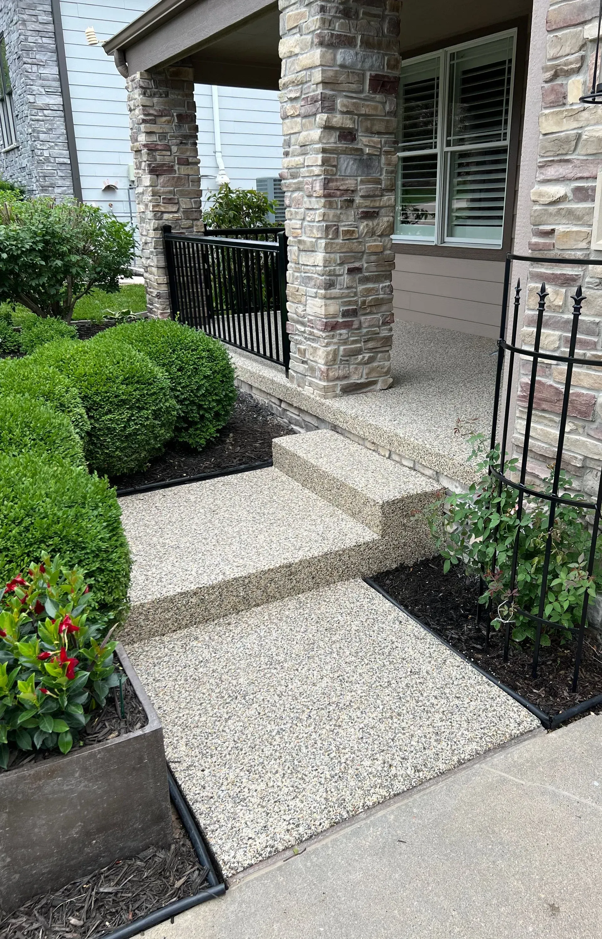 A stone-textured porch with steps, surrounded by green shrubs, dark mulch, and a metal trellis, leading to a home entrance.