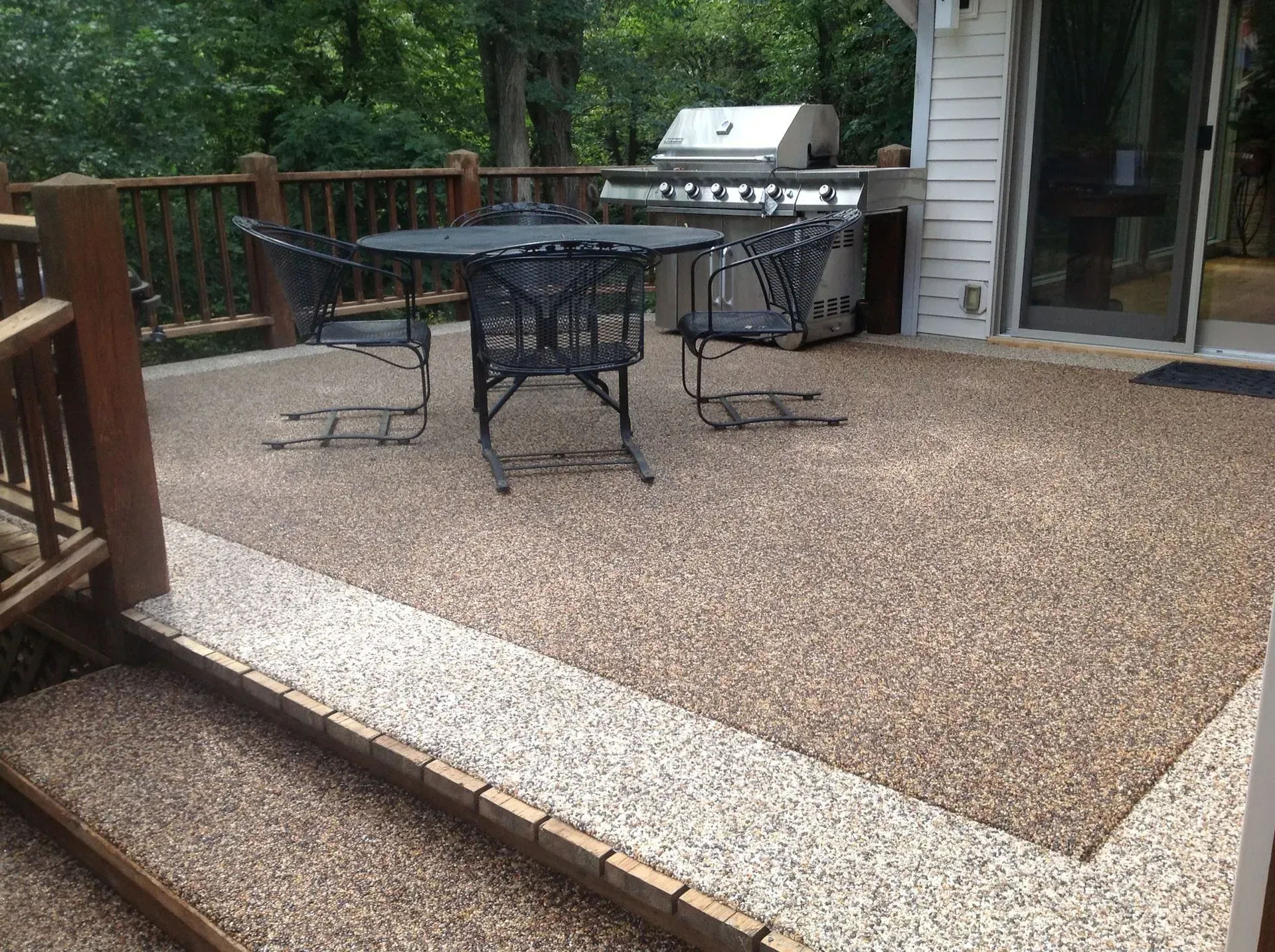 A outdoor patio deck with pebble-texture flooring, a metal table and chairs, and a built-in grill near a house exterior.