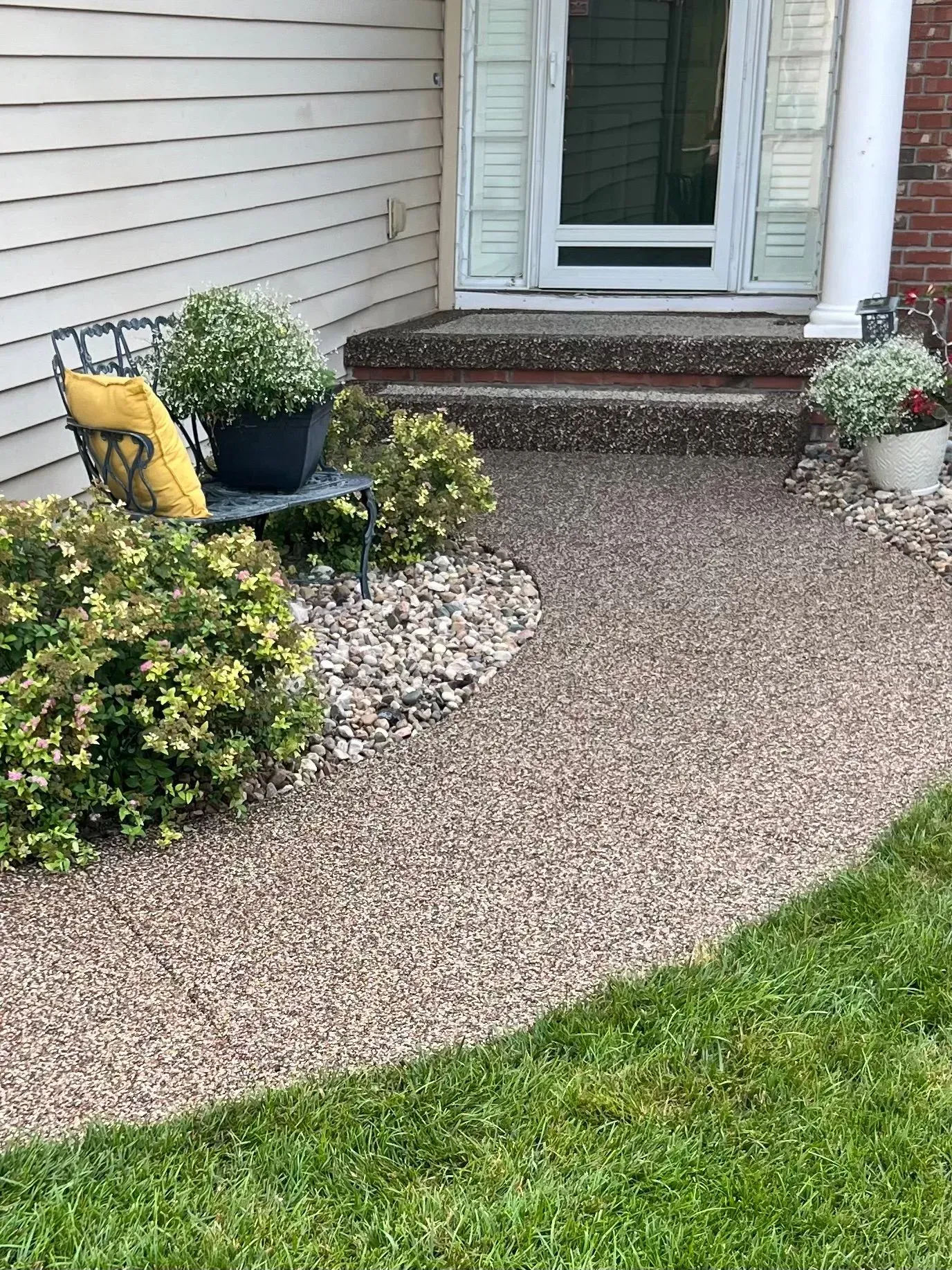 A landscaped front walkway with pebble surfacing, shrubs, and a metal bench with a yellow cushion leading to front steps.