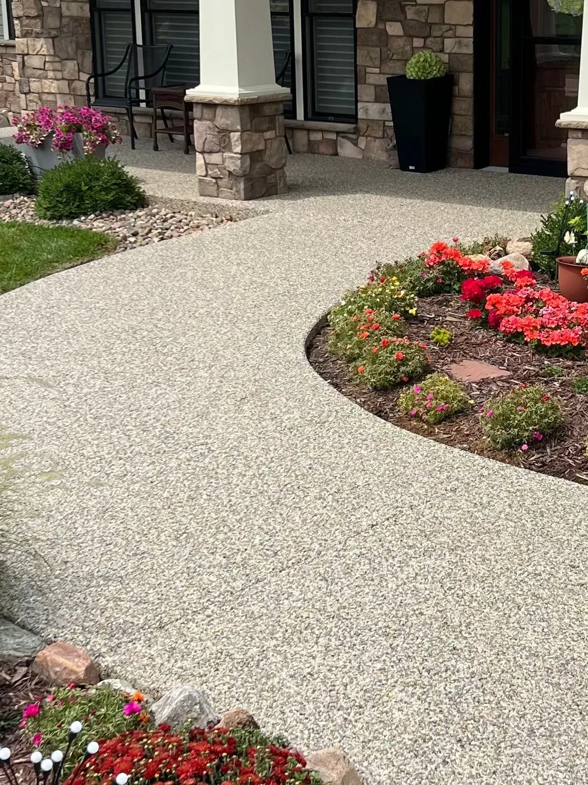 A curved, aggregate concrete walkway leads toward the entrance of a stone house with landscaped flower beds.