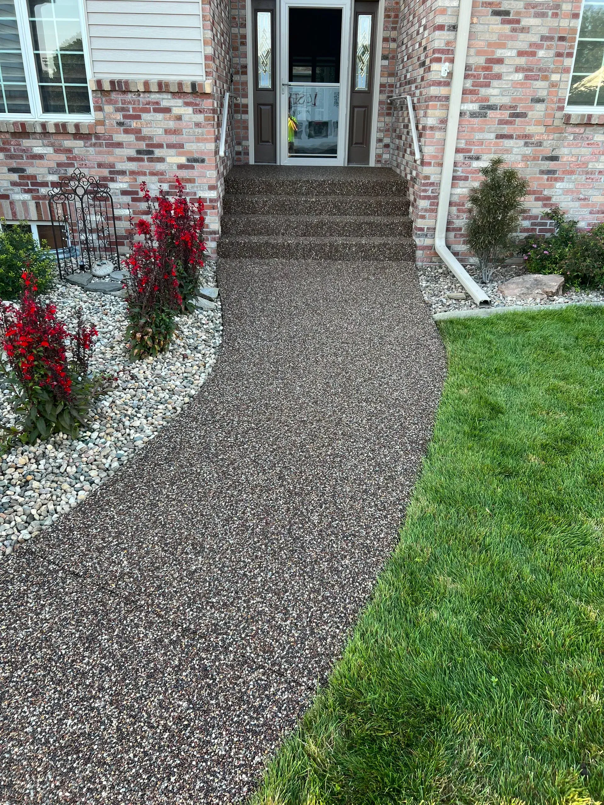 A brick house entrance with a brown stone-paved walkway, bordered by decorative red shrubs and a green lawn.