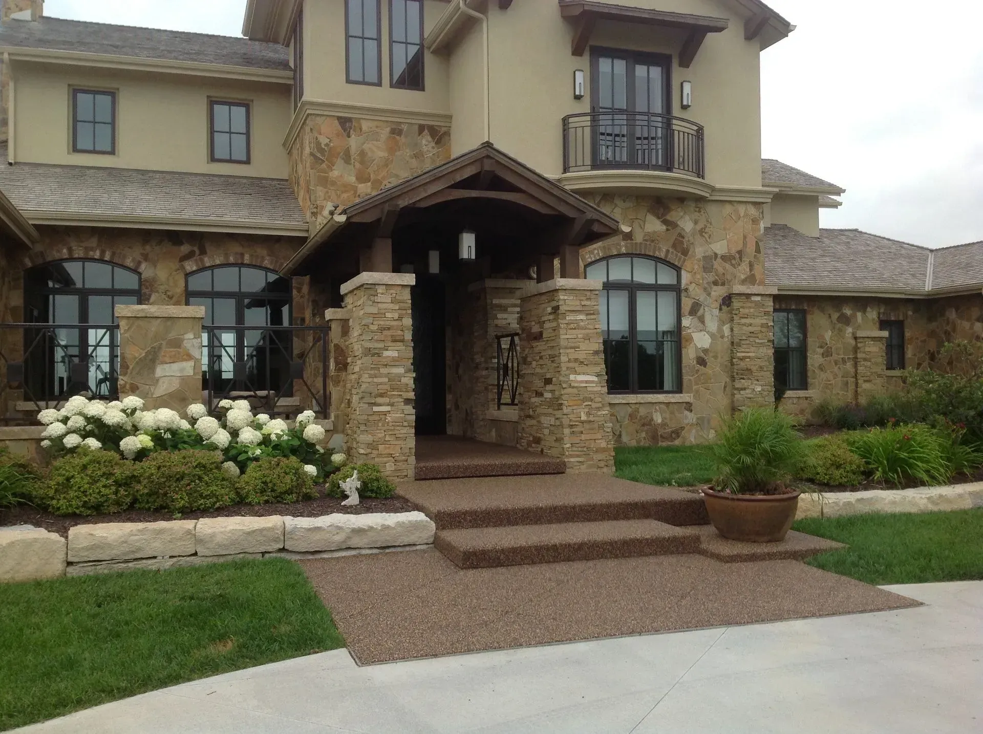 A two-story stone house exterior featuring an arched entrance, concrete steps, a small balcony, and landscaped shrubbery.