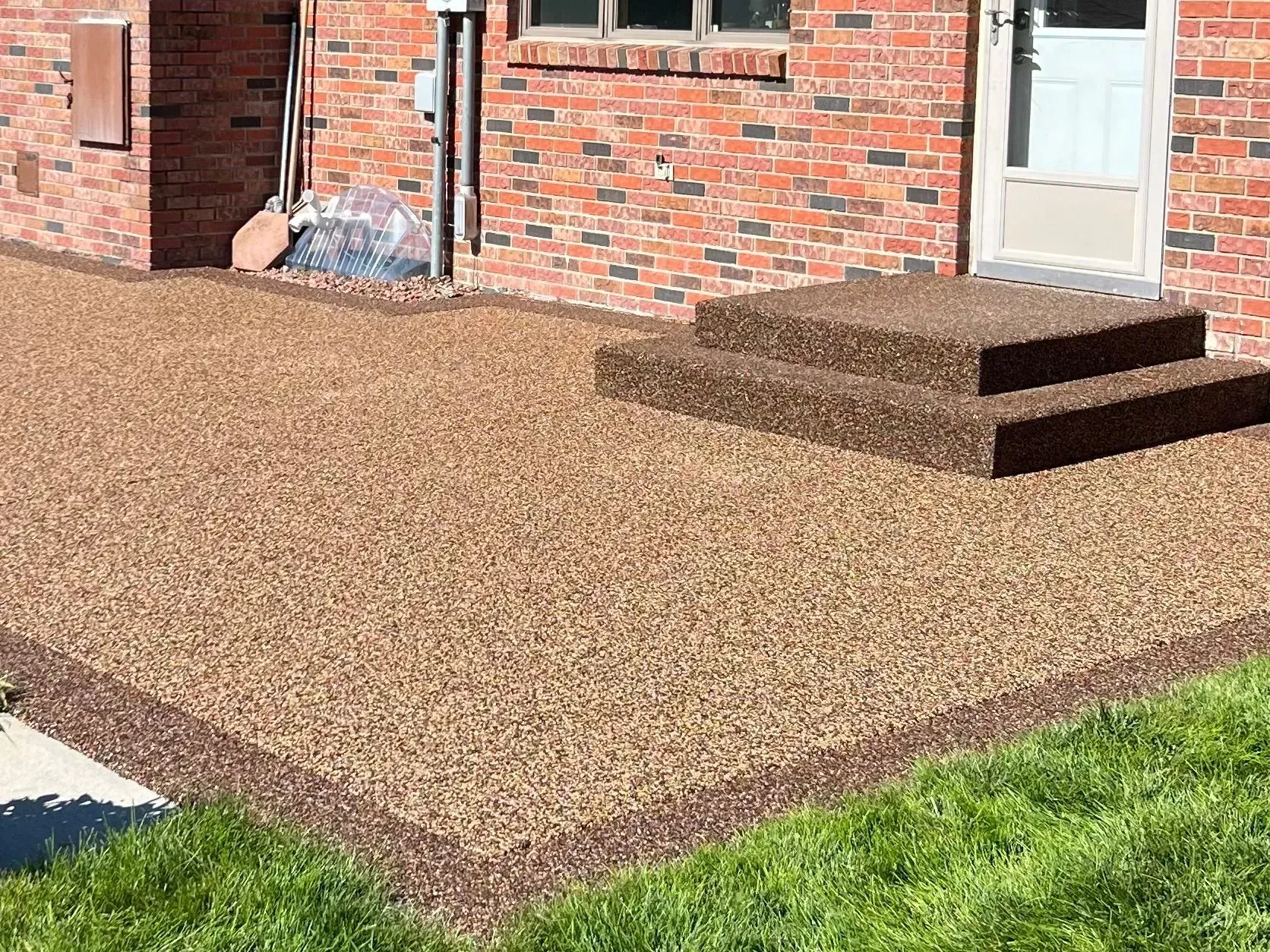 A tan pebble-textured patio with a matching two-step landing leading to a white door against a brick house exterior.