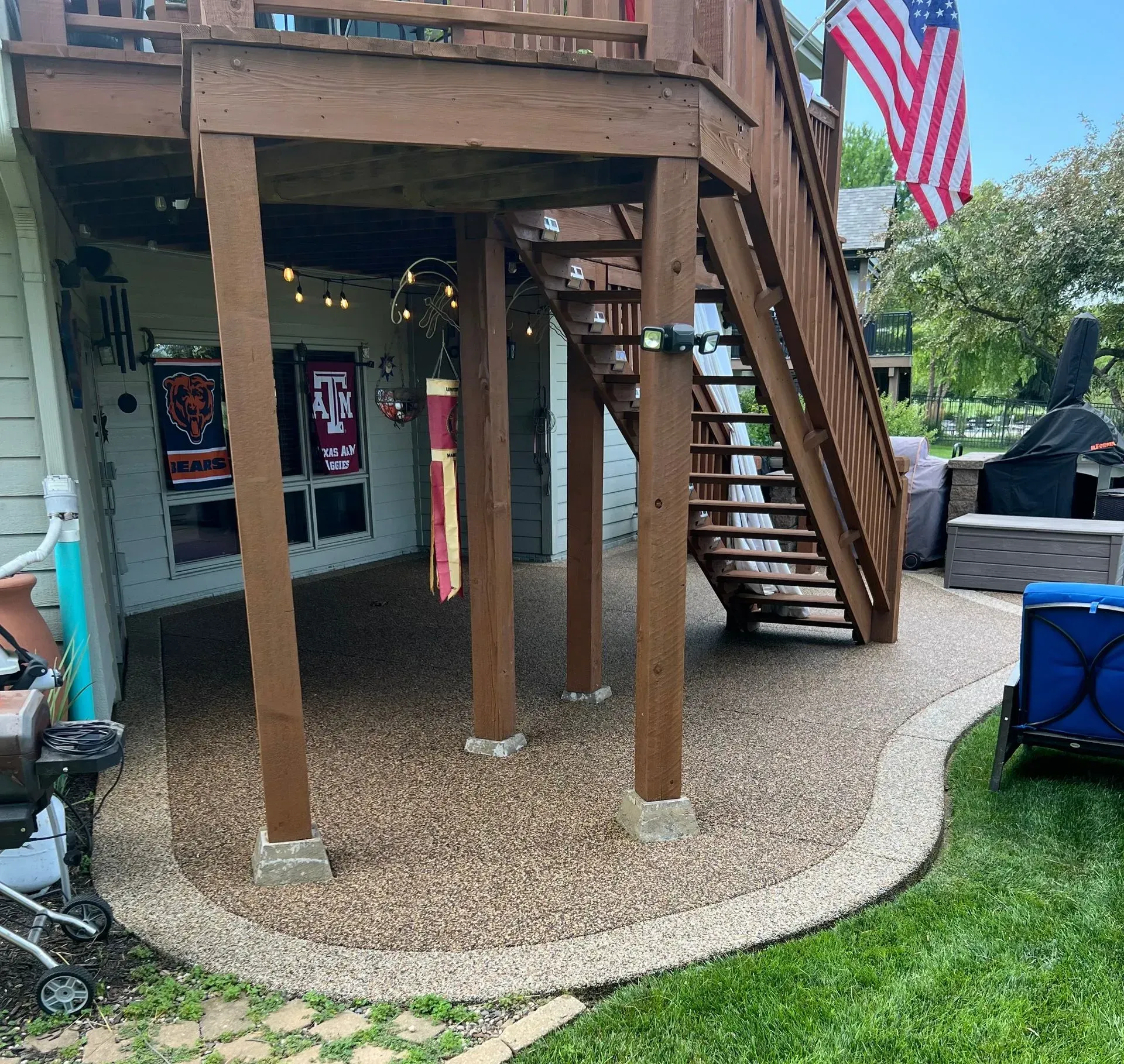 A wooden deck with a staircase overhead, a gravel patio area, and an American flag hanging from the side of the stairs.