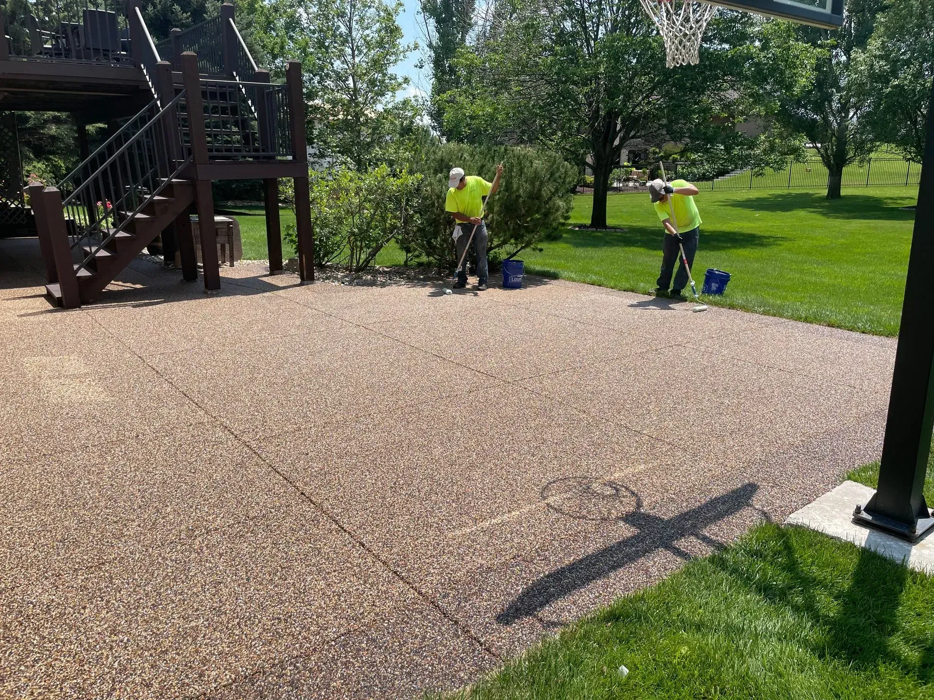 Two workers in high-visibility yellow shirts finish a pebble-texture patio near a wooden deck and basketball hoop.