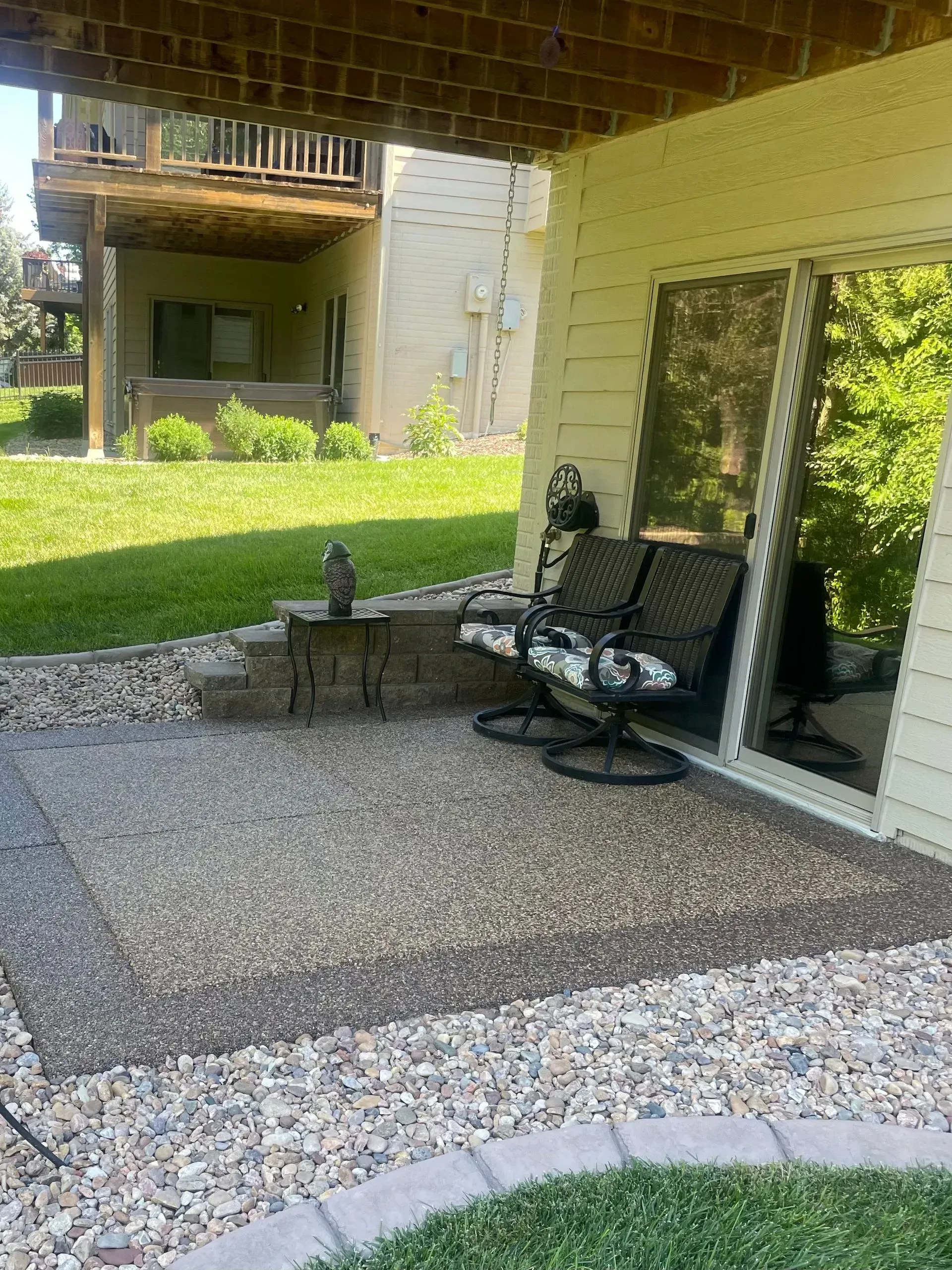 A covered gravel patio with two patio chairs and a small table next to a sliding glass door, facing a grassy yard.