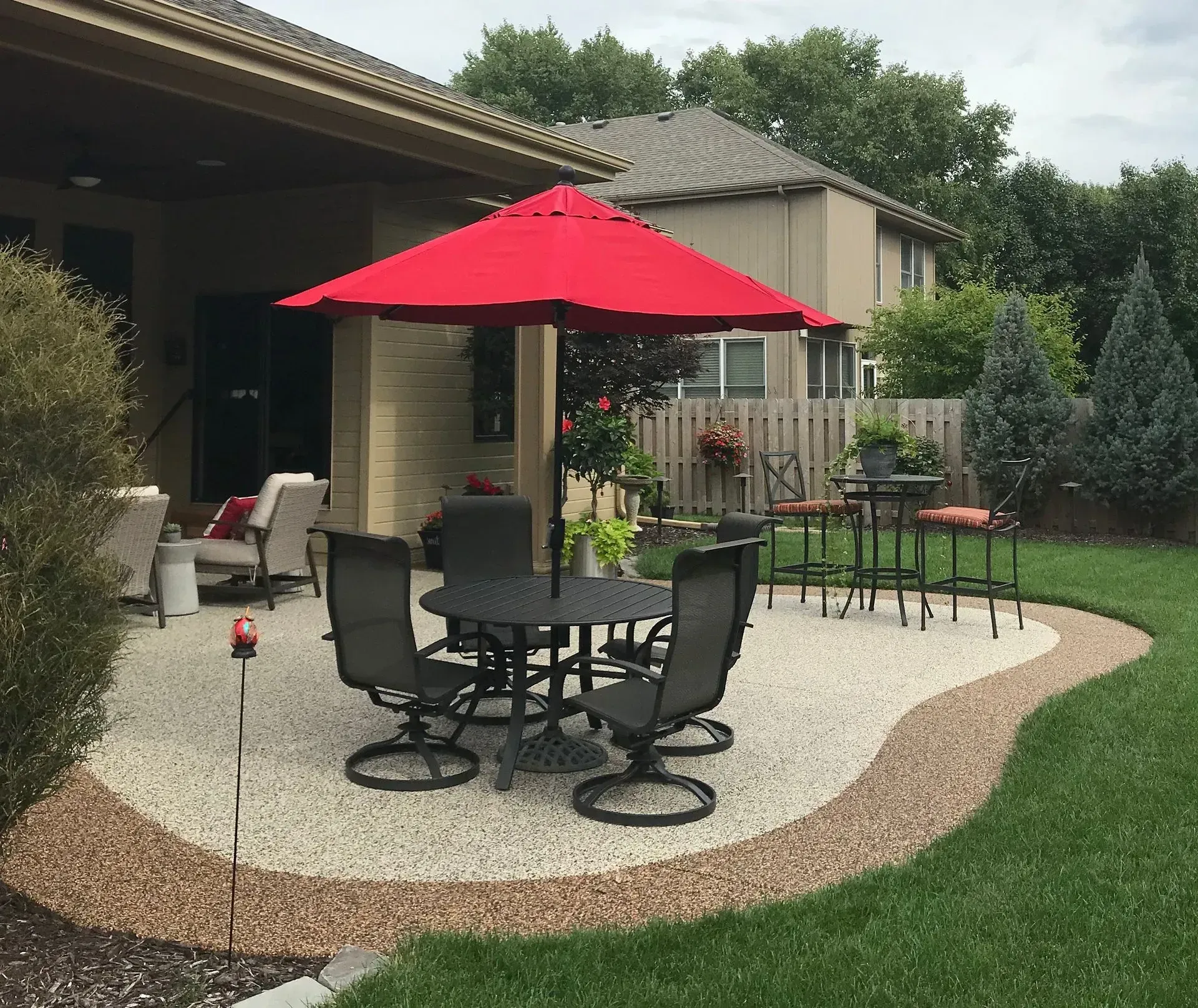 A red patio umbrella centered over a dining table and chairs on a gravel patio beside a house and grassy lawn.