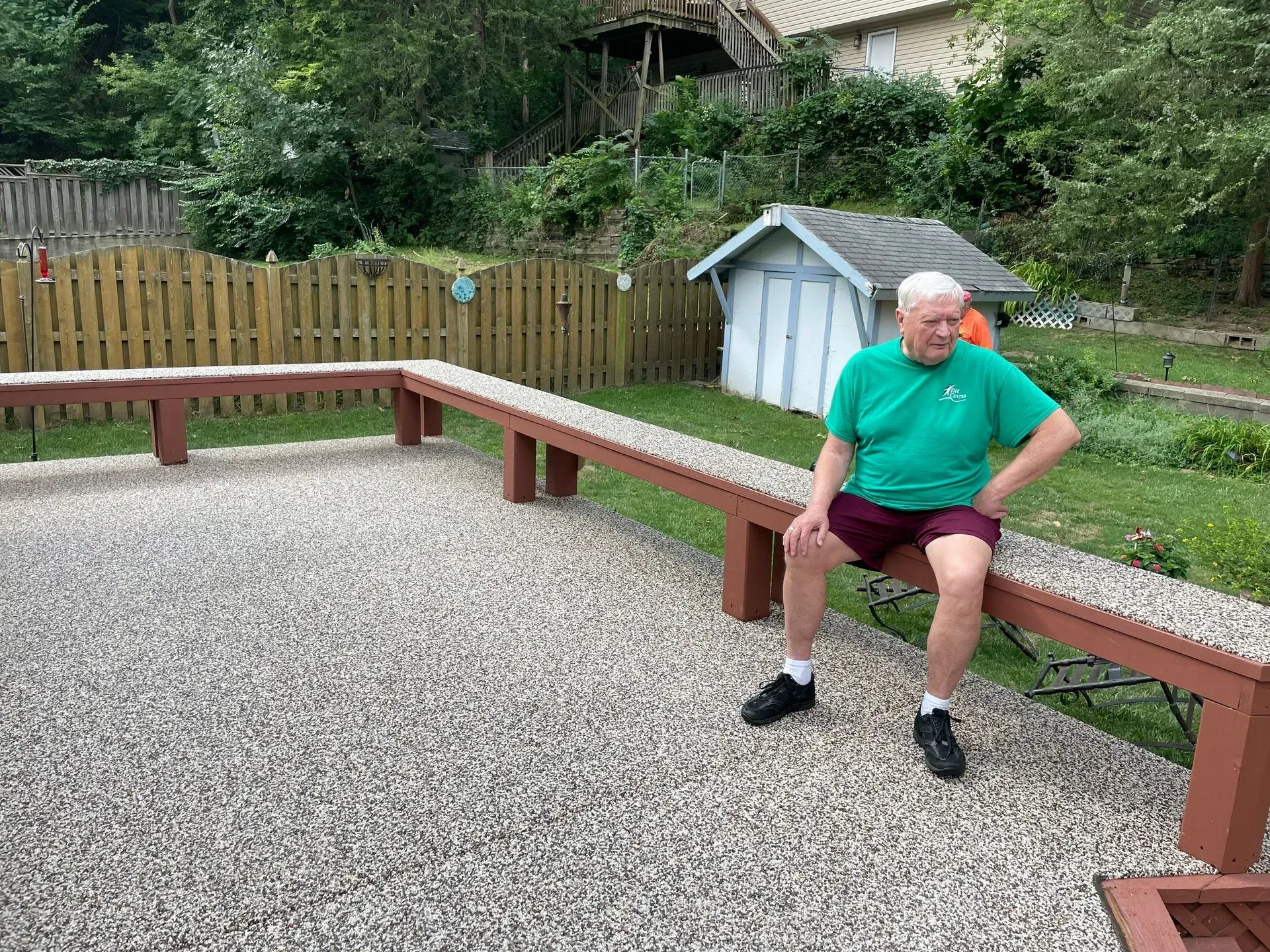 A person in a green shirt and maroon shorts sits on a long, U-shaped wooden bench surrounding a gravel patio.
