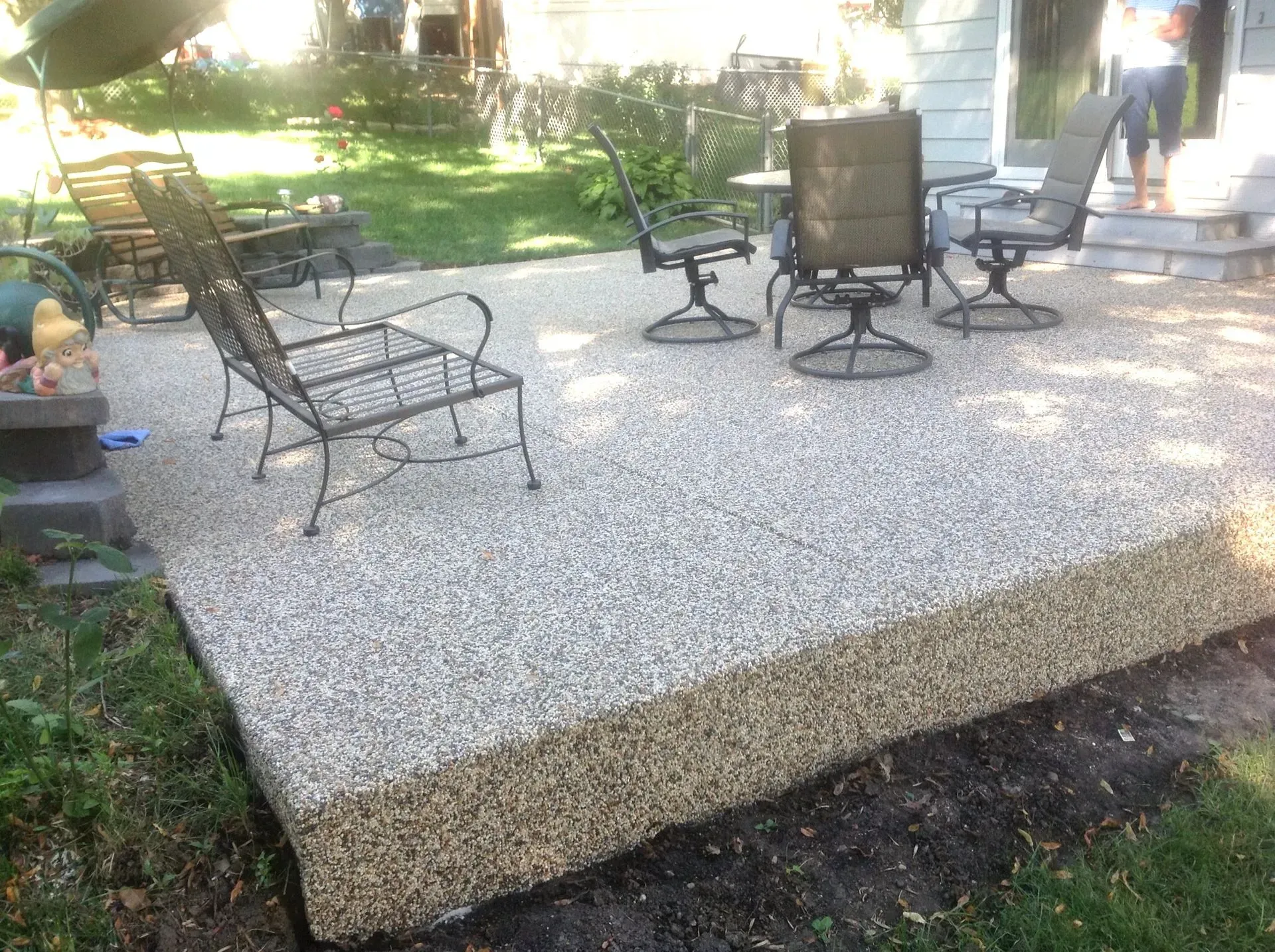 A backyard patio made of exposed aggregate concrete, furnished with metal chairs and a table on a sunny day.