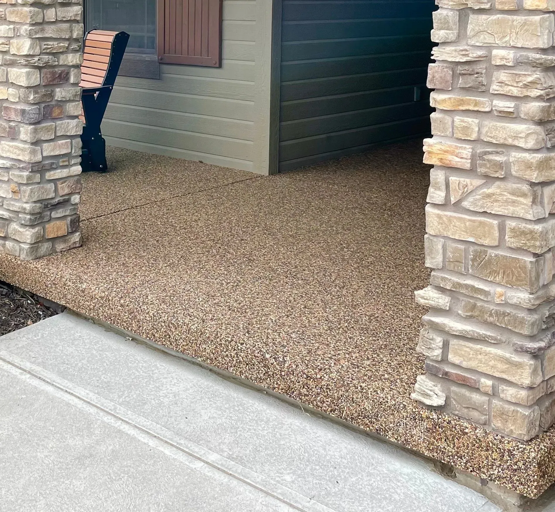 A porch featuring a textured, light brown pebble-stone floor framed by two stone columns and olive-colored siding.