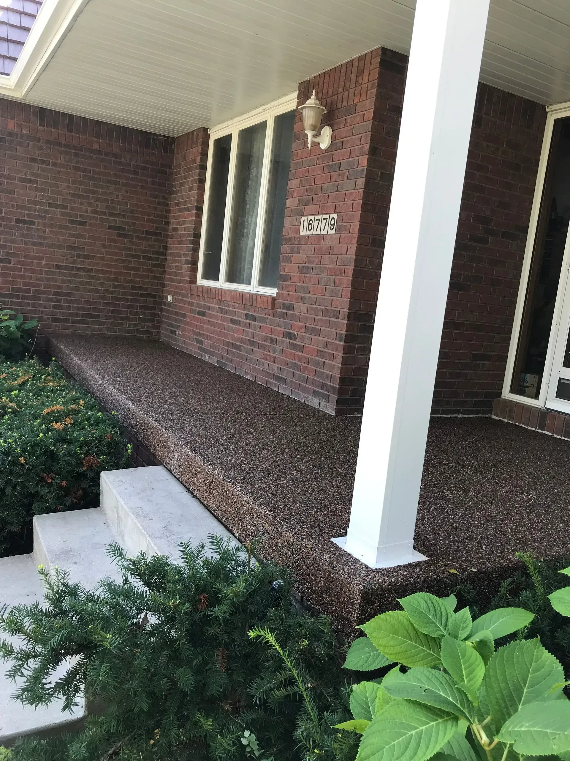 A dark, textured stone porch entrance to a brick house with a white support pillar, concrete steps, and green shrubs.