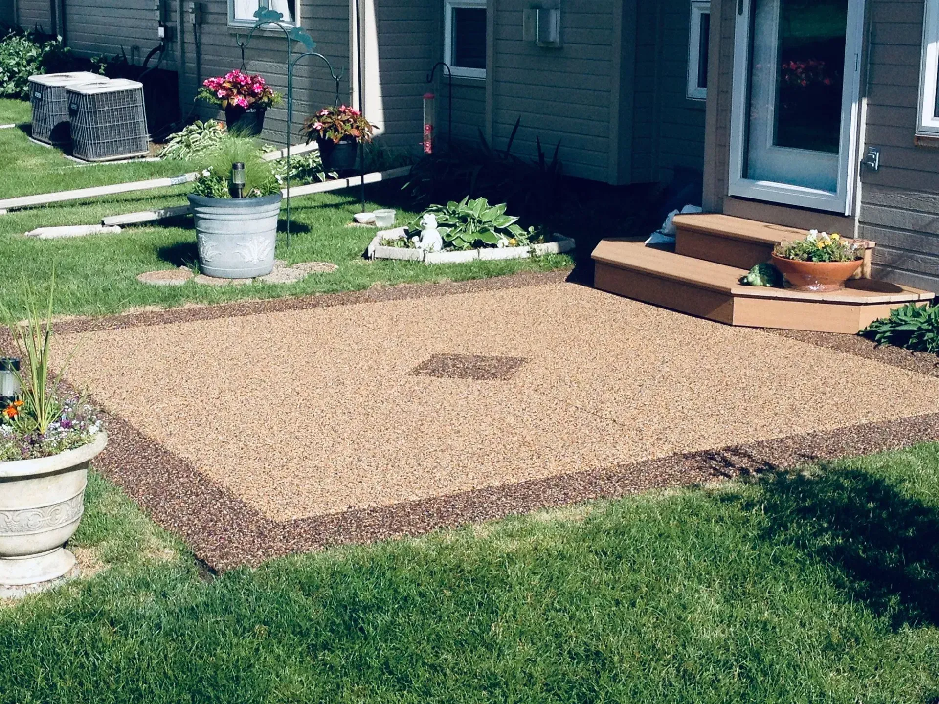 A square patio made of light brown gravel with a dark stone border, located in a grassy yard by a house entrance.
