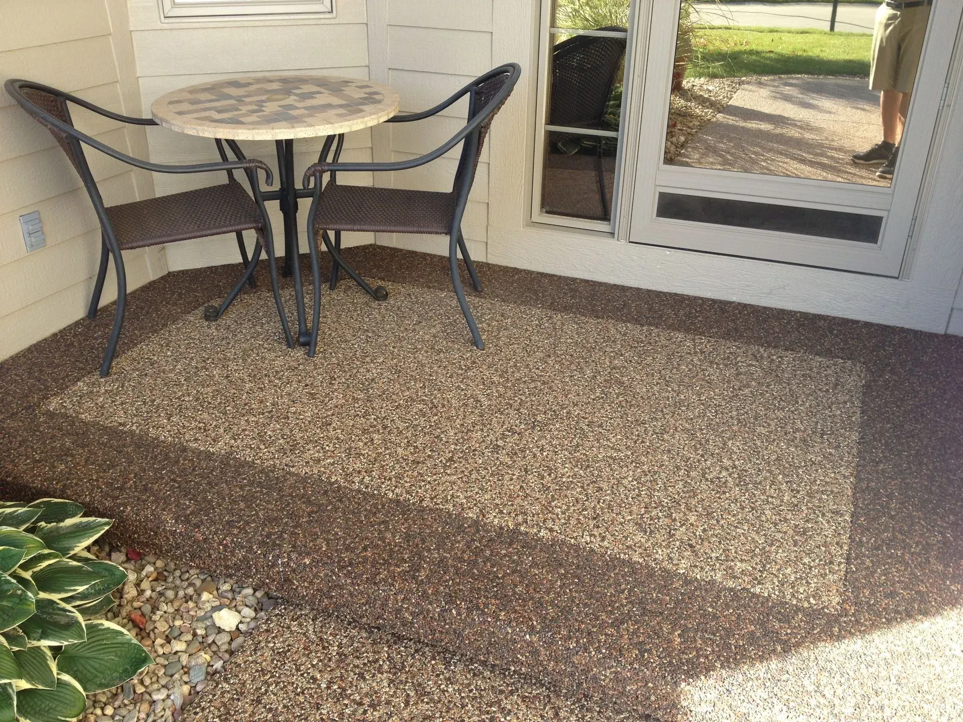 A small stone-textured patio with a bistro table and two chairs, situated in a corner outside a glass door.