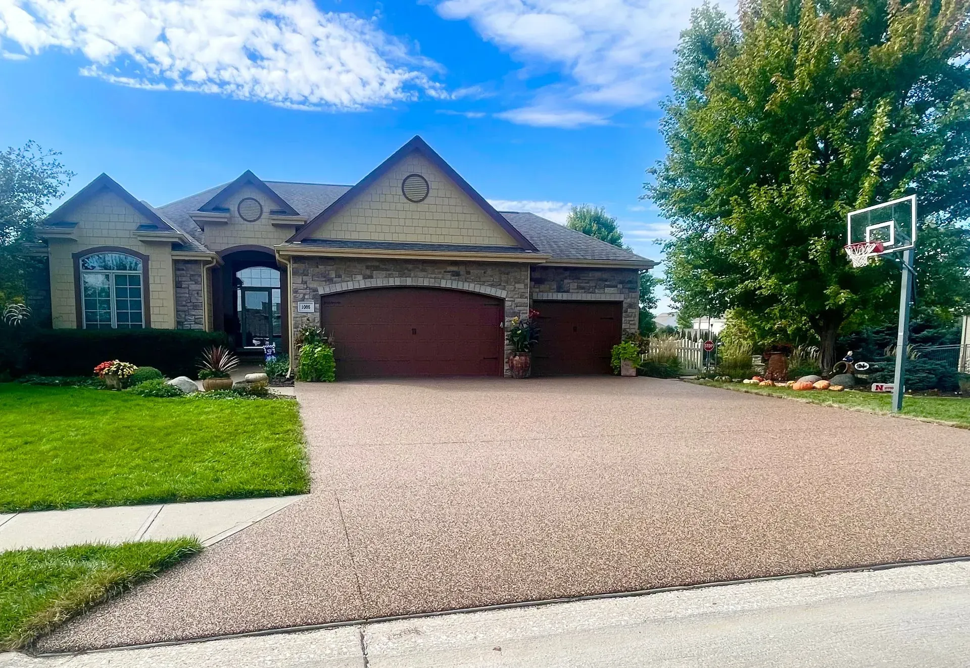 A tan, multi-gabled suburban home with a dark brown three-car garage and a large gravel driveway under a blue sky.