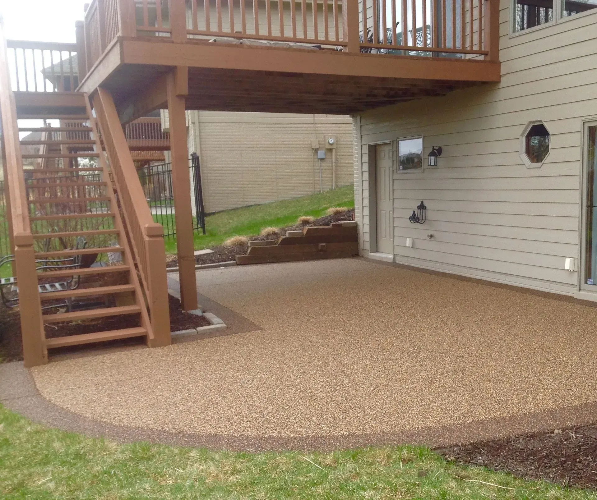 A beige stone-textured patio sits beneath a wooden deck with stairs in a backyard setting.