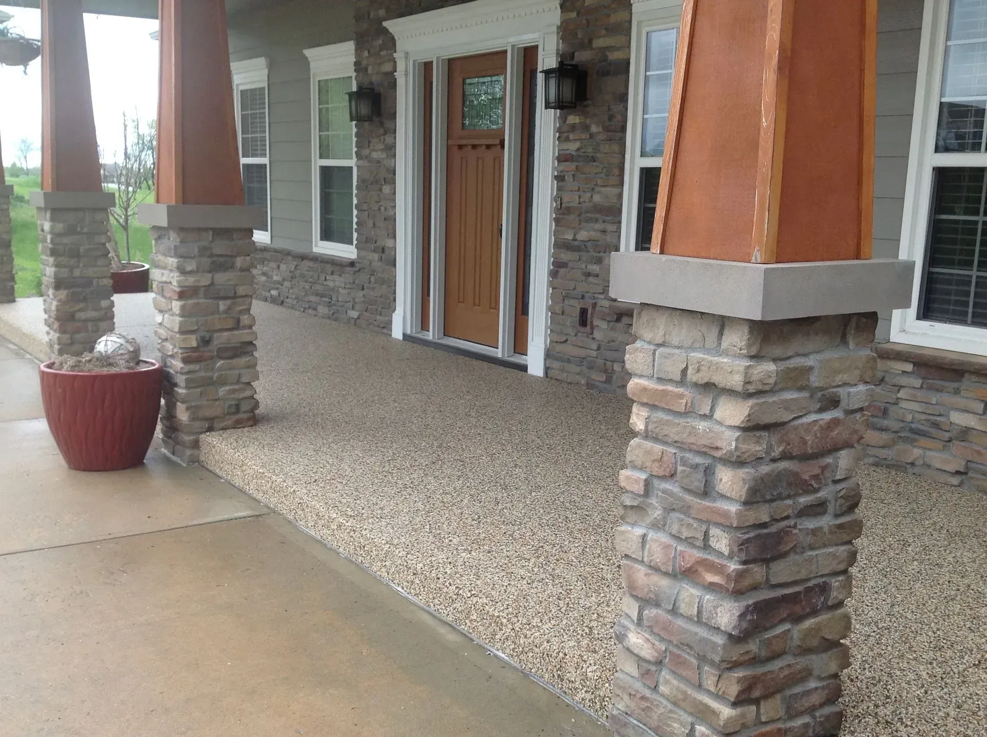 A covered porch with stone pillar bases, wooden columns, a central wooden door, and a textured, pebbled floor surface.