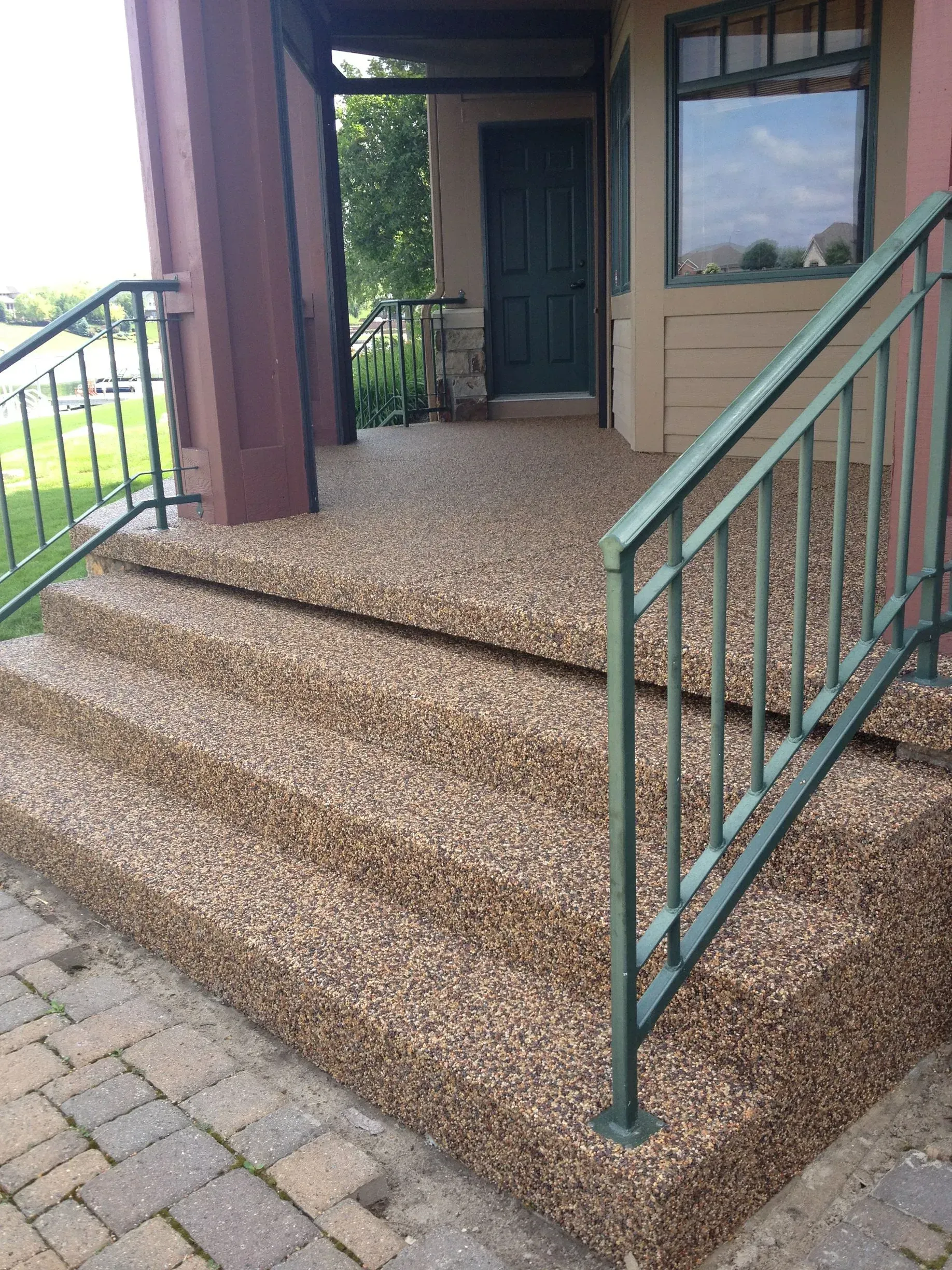Concrete steps and a porch covered in textured brown pebble surfacing, featuring green metal railings.