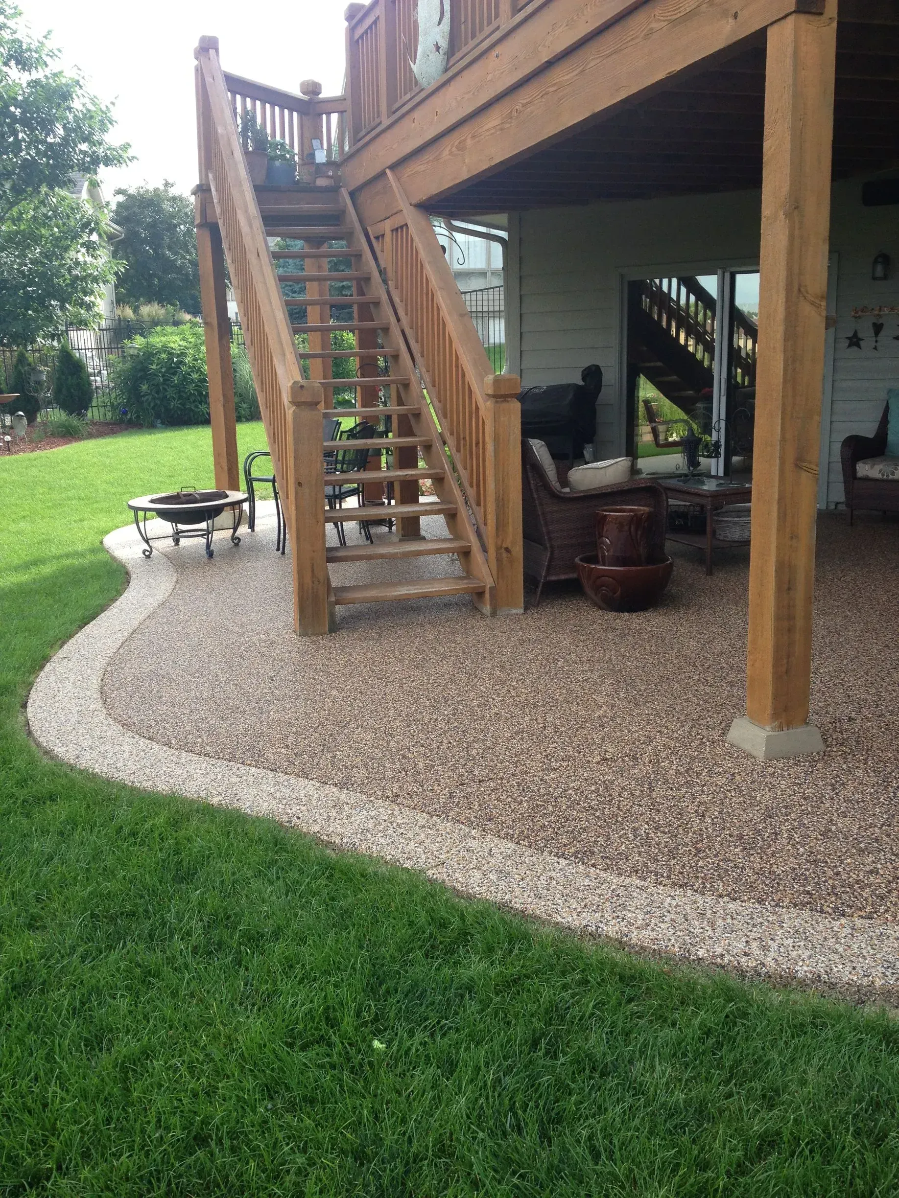 A wooden deck staircase overlooks a patio made of brown gravel with a lighter stone border, set against a green lawn.