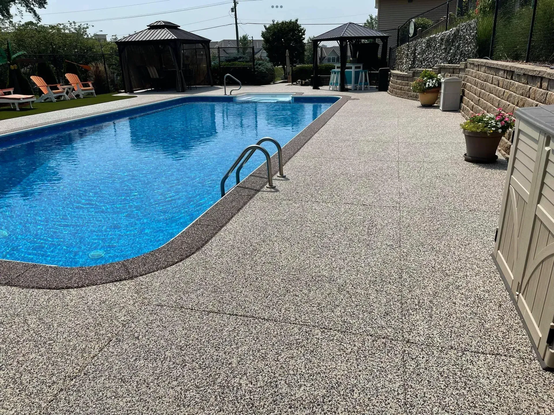 A bright blue swimming pool with a speckled stone deck, surrounding gazebos, and potted plants on a sunny day.