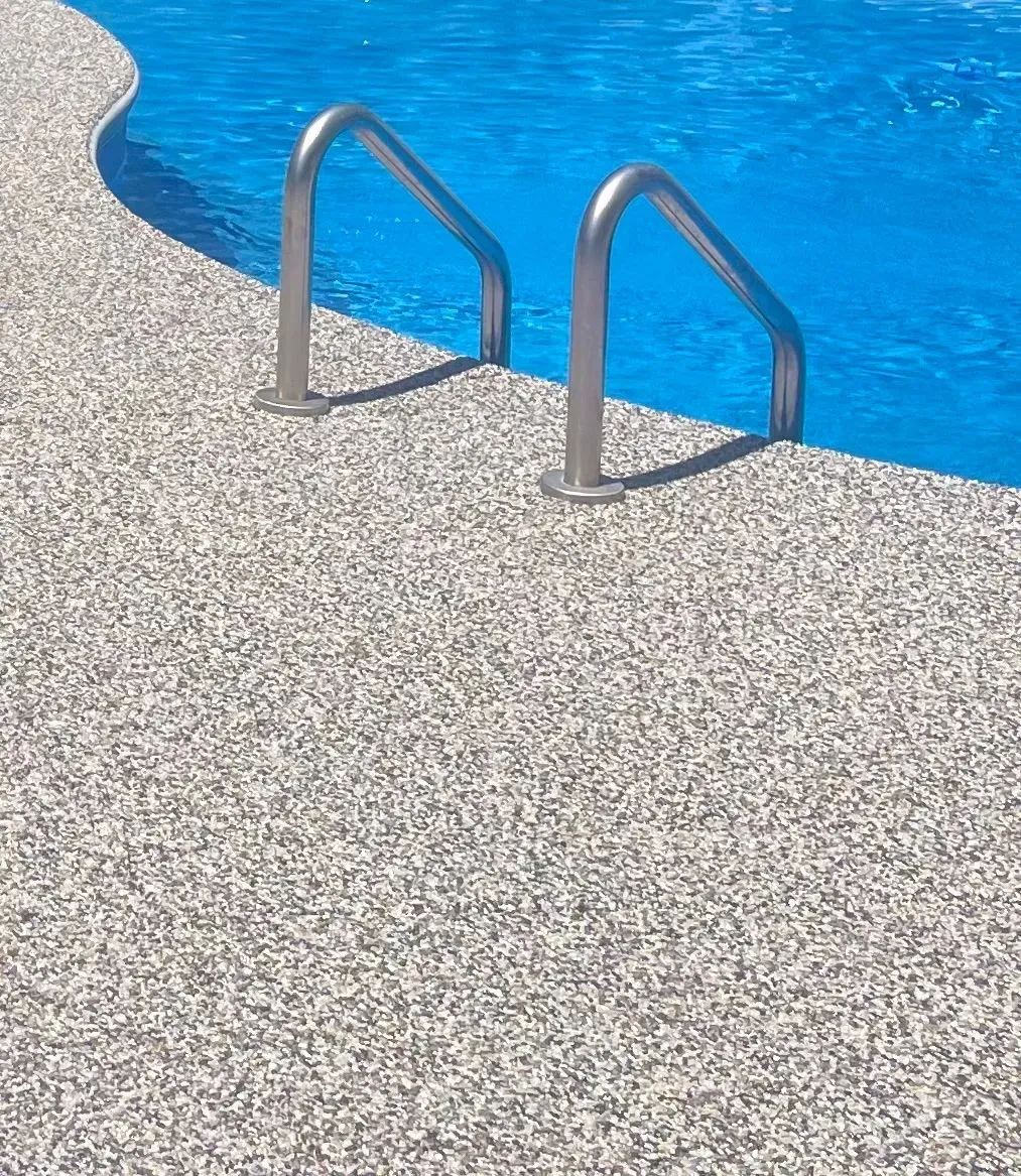 A stainless steel pool ladder anchored to a textured gray and white stone pool deck next to bright blue water.