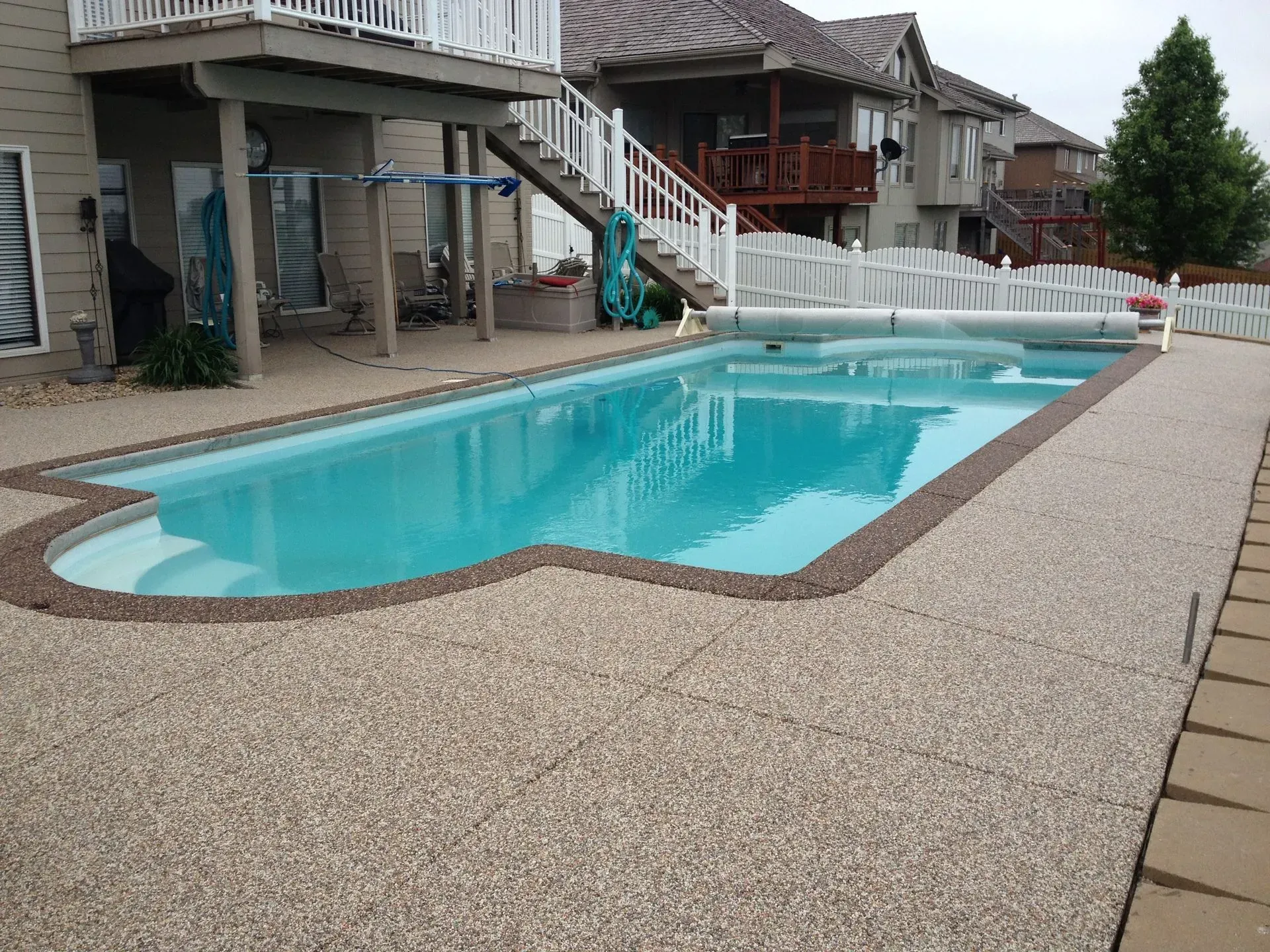 A rectangular backyard swimming pool with a stone deck, adjacent to a house with an elevated wooden deck and stairs.