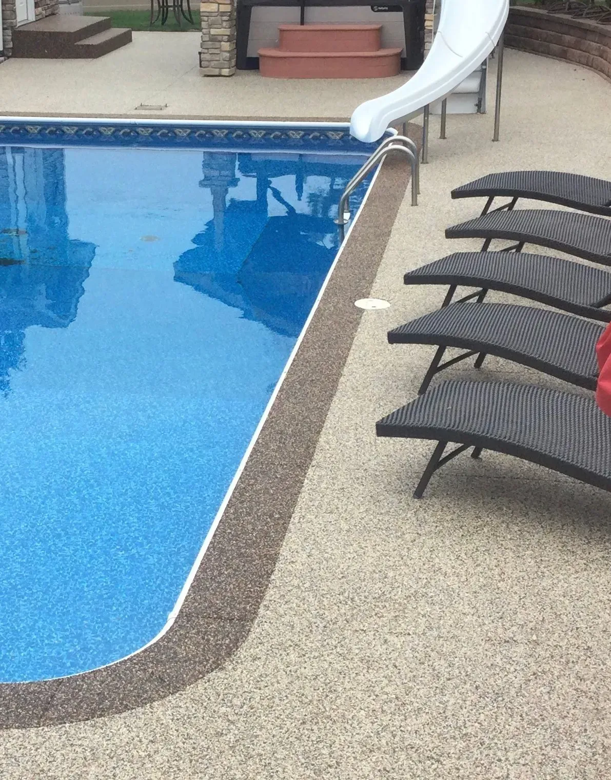 A row of five dark wicker lounge chairs sits on a pebble-textured pool deck next to a blue swimming pool with a slide.