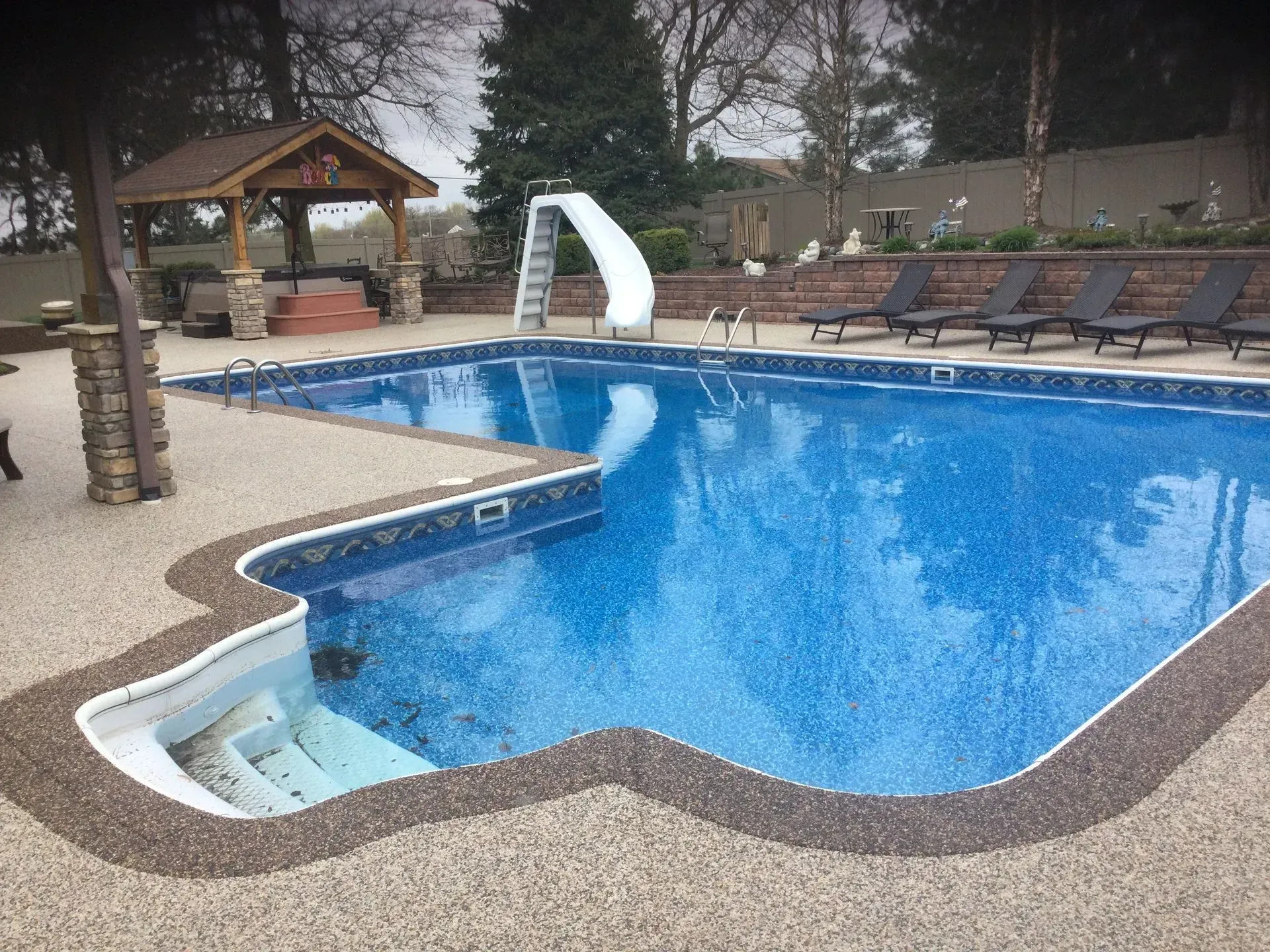 An L-shaped swimming pool with blue water and white steps, featuring a slide and a shaded gazebo on a stone patio.