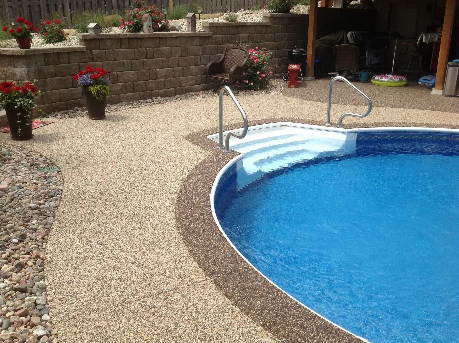 A backyard swimming pool with blue water and white steps, surrounded by a light-colored pebbled patio and retaining wall.