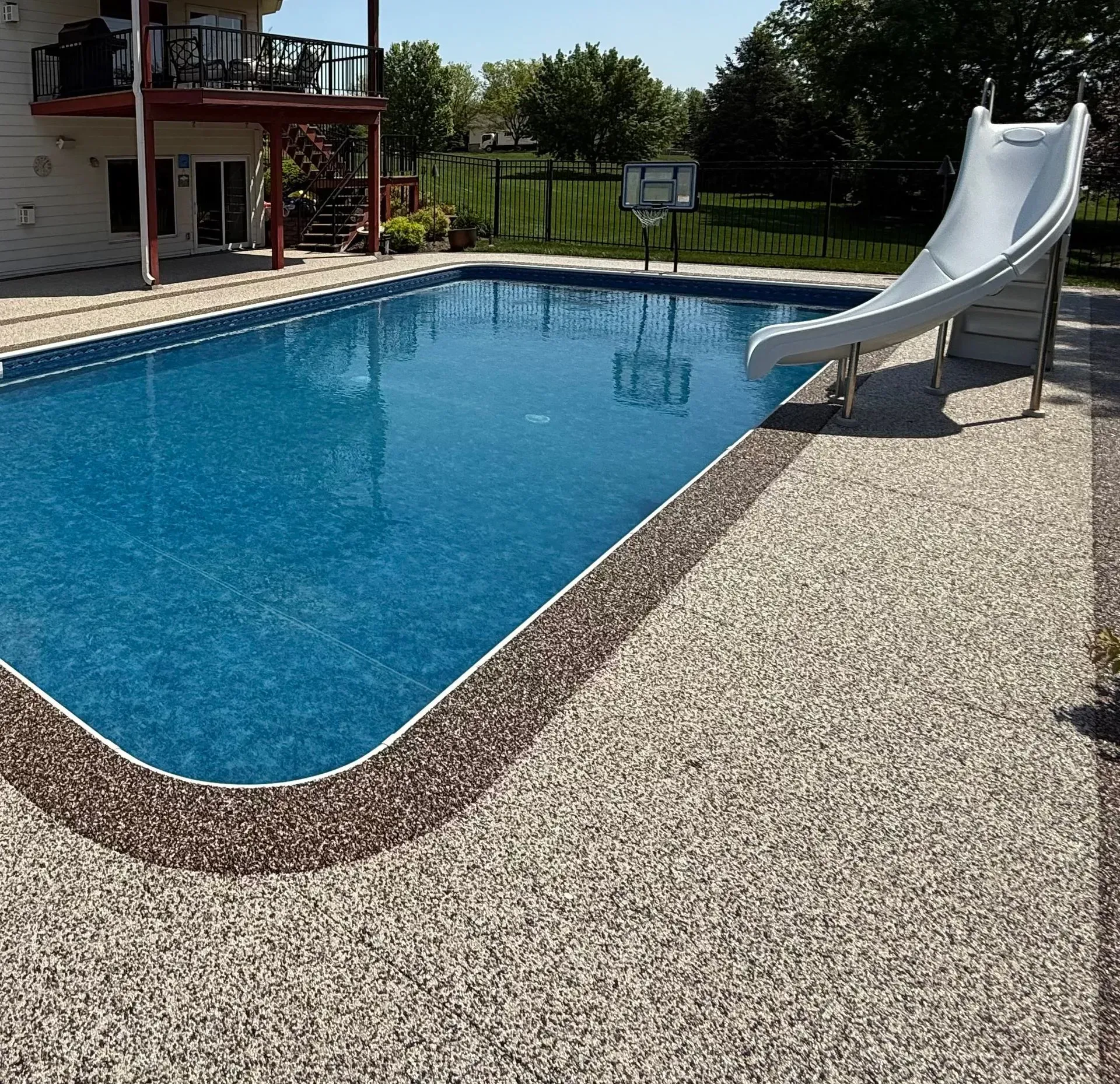 A bright blue rectangular swimming pool with a white slide and basketball hoop, set against a house and yard.