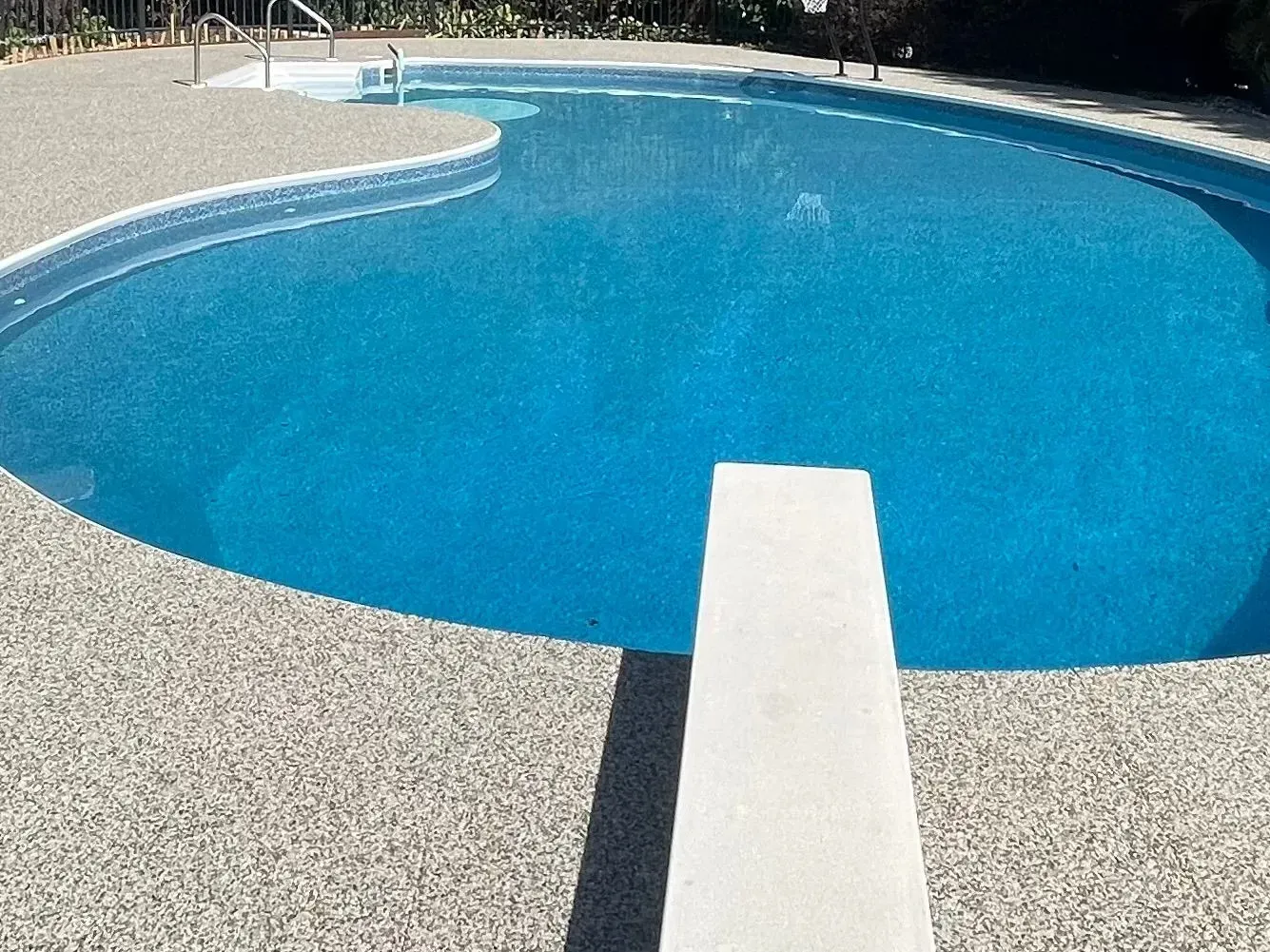 A blue, kidney-shaped swimming pool with a diving board in the foreground and a textured concrete deck surrounding it.