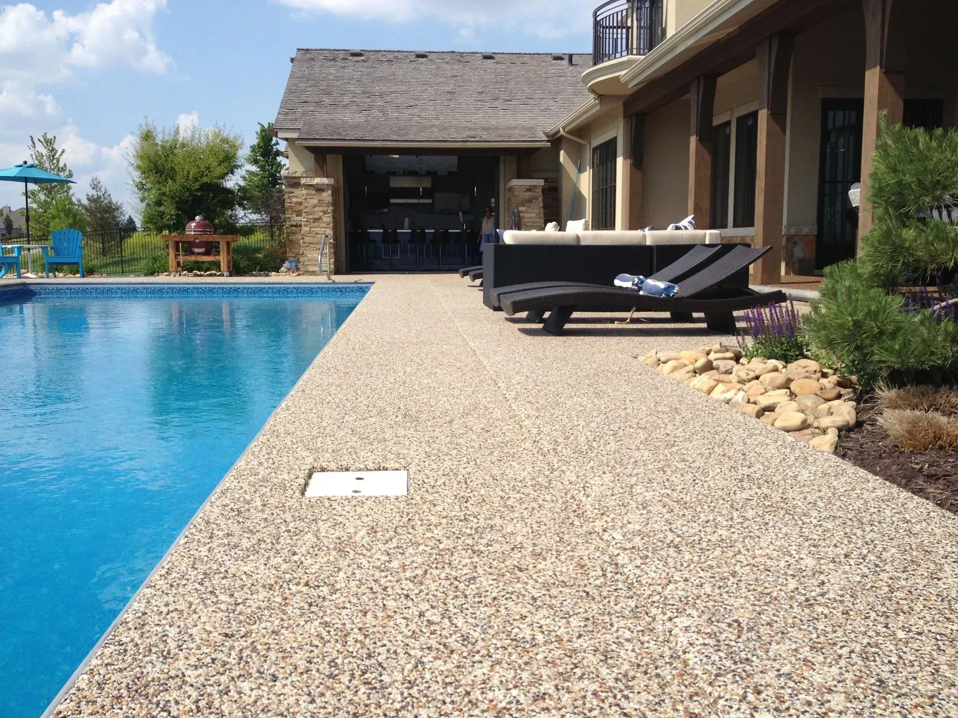 A swimming pool deck made of textured, light-colored aggregate, with lounge chairs and a house patio in the background.
