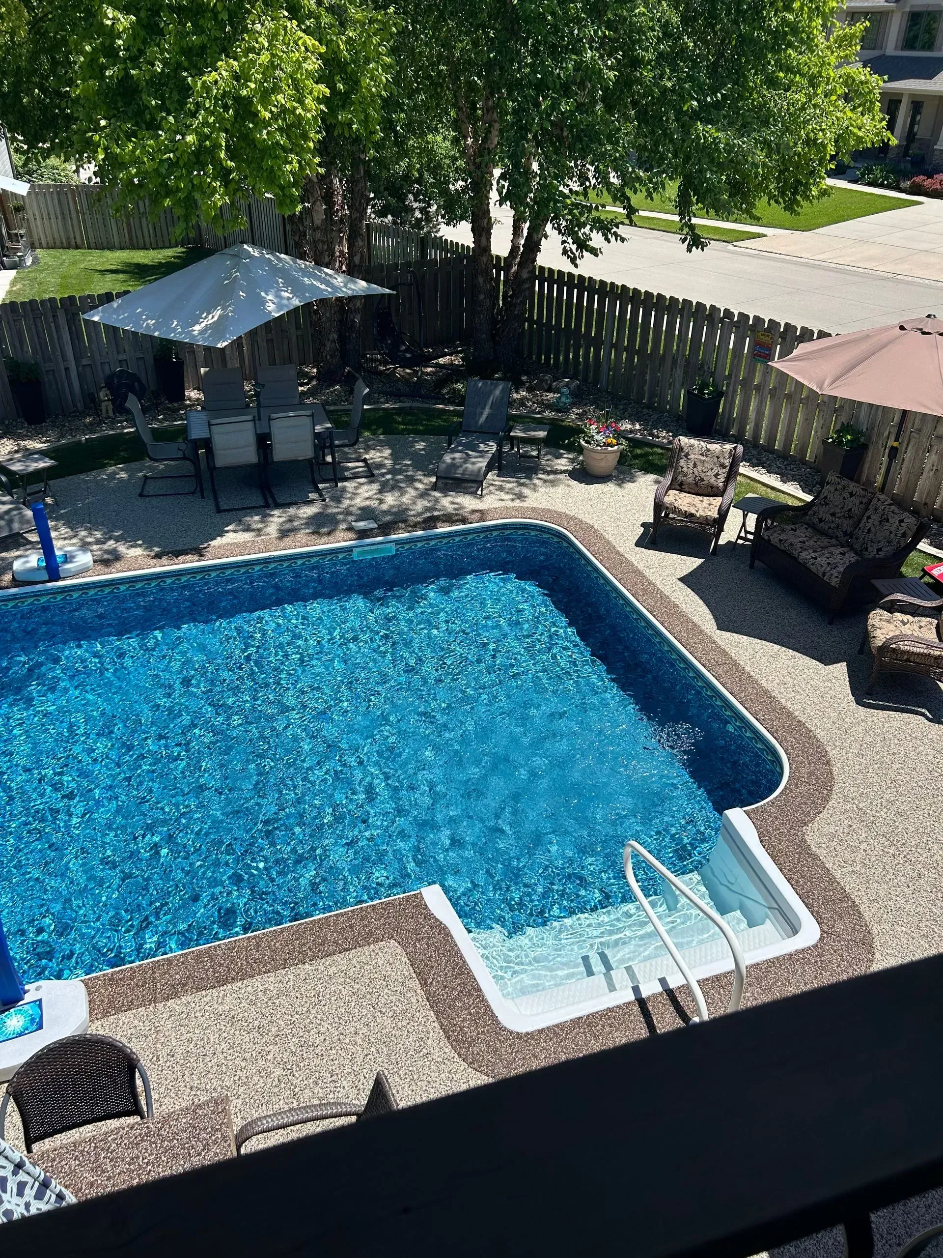 A high-angle view of a backyard swimming pool surrounded by a gravel patio with lounge chairs and patio umbrellas.