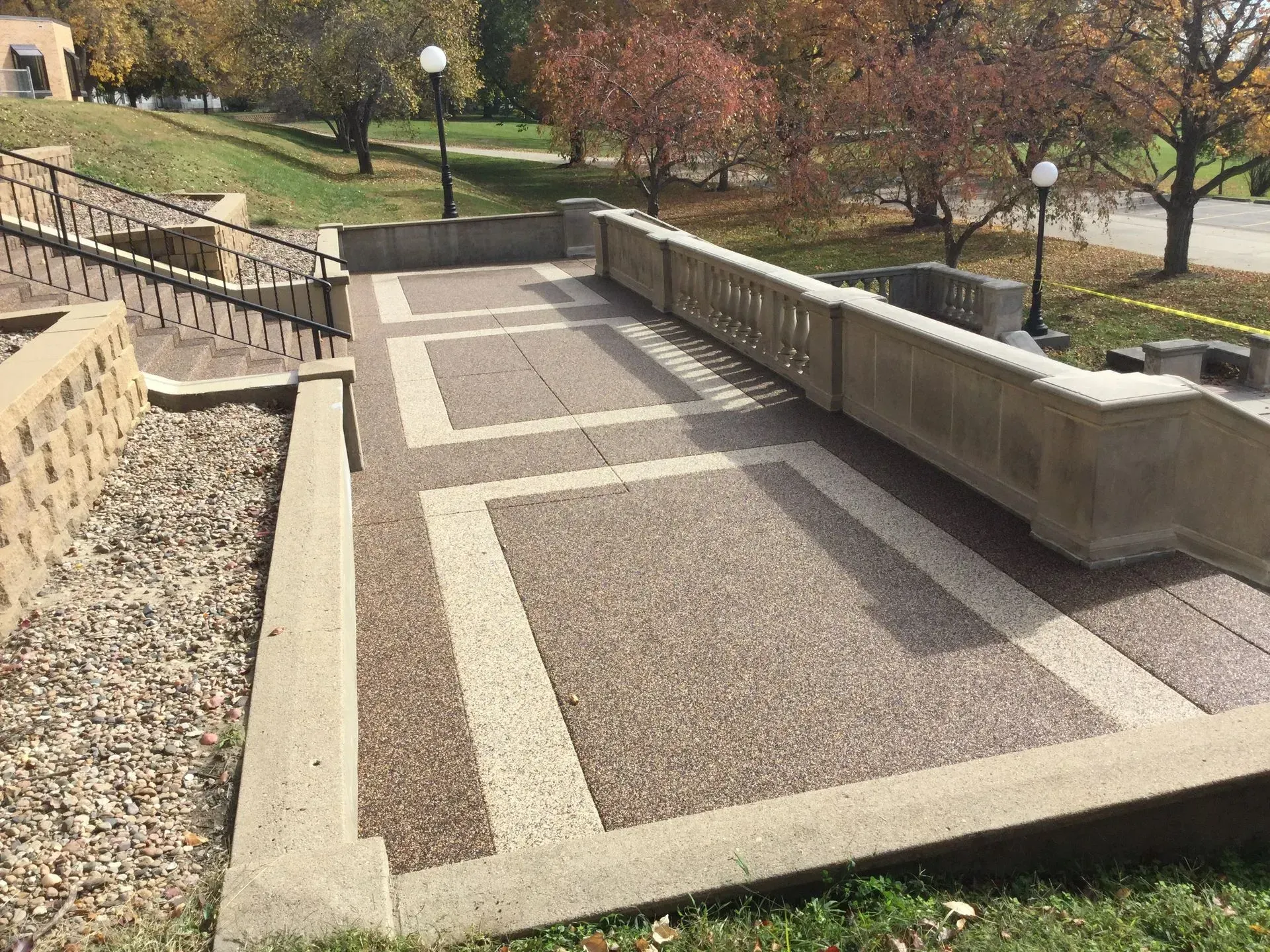 A wide outdoor concrete staircase with textured landings and stone railings, set in a park-like landscape during autumn.