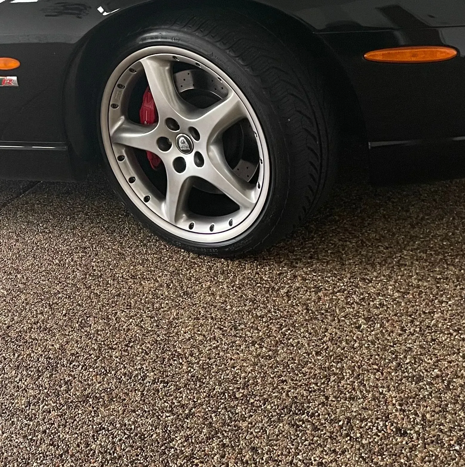 A close-up of a black sports car's silver alloy wheel and orange side marker on a textured, speckled garage floor.