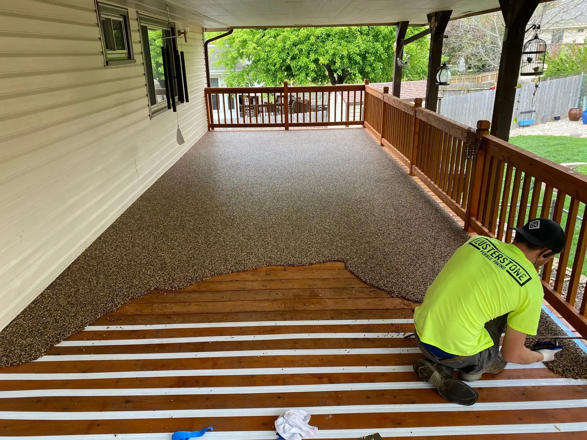 A worker in a neon yellow shirt applies a textured stone coating to part of a wooden deck, leaving some boards exposed.