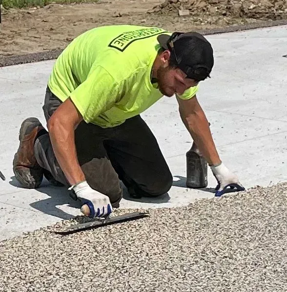 A worker in a neon green shirt kneels on a concrete surface, using a hand trowel to smooth out a gravel edge.