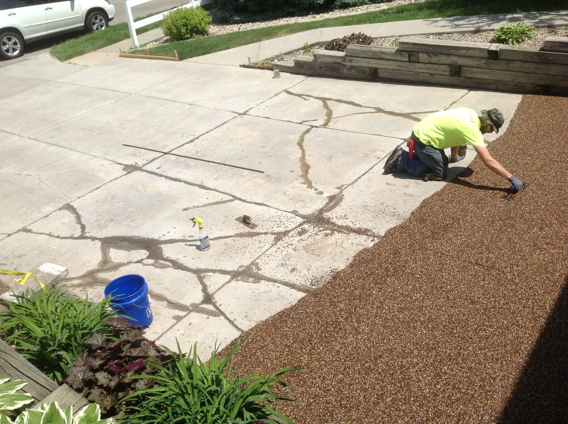 A person in a neon yellow shirt kneeling on a cracked concrete driveway, working on the surface next to brown mulch.
